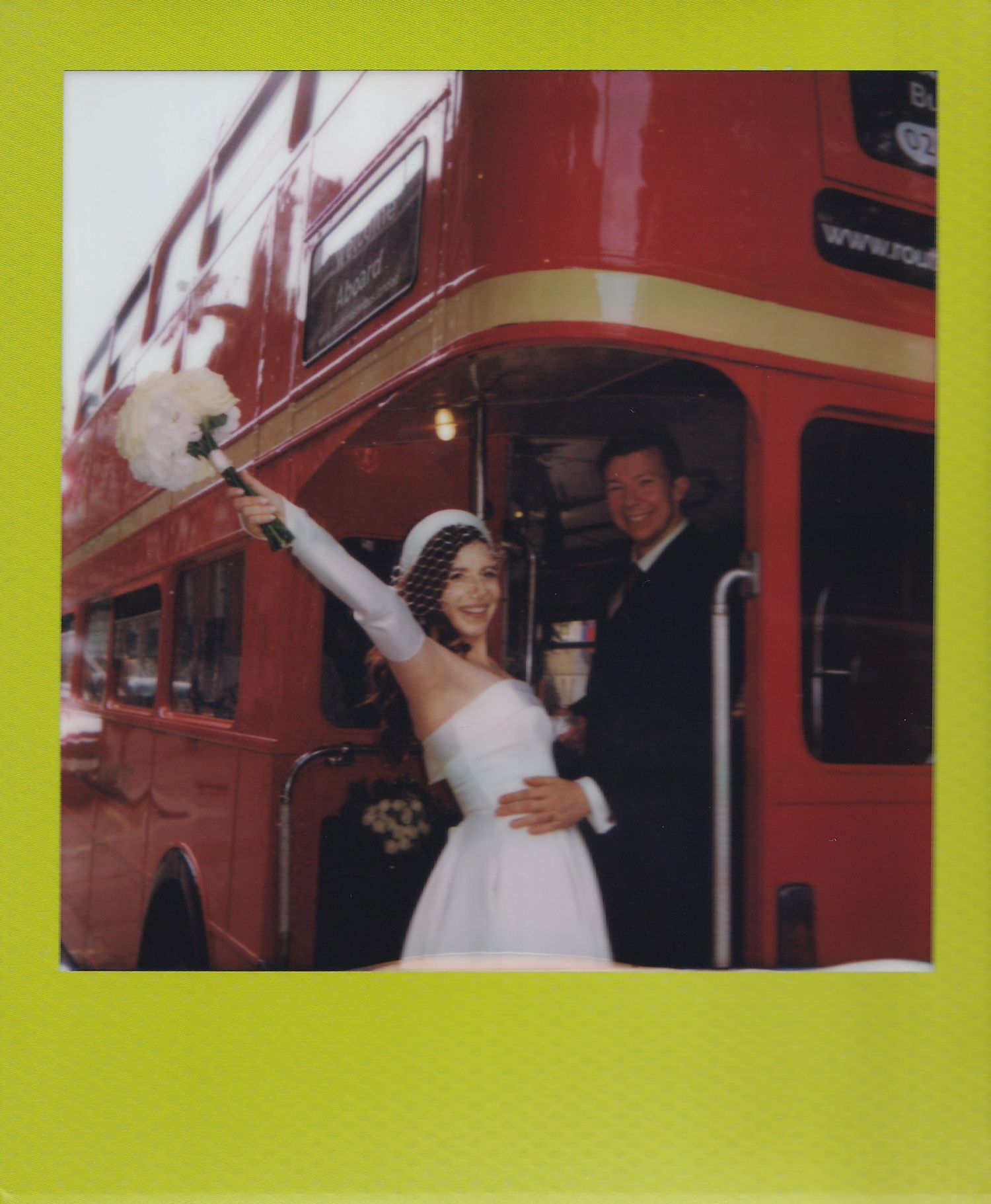 A bride and groom pose joyfully in front of a red double-decker bus, bride holding bouquet aloft in a Polaroid photo.