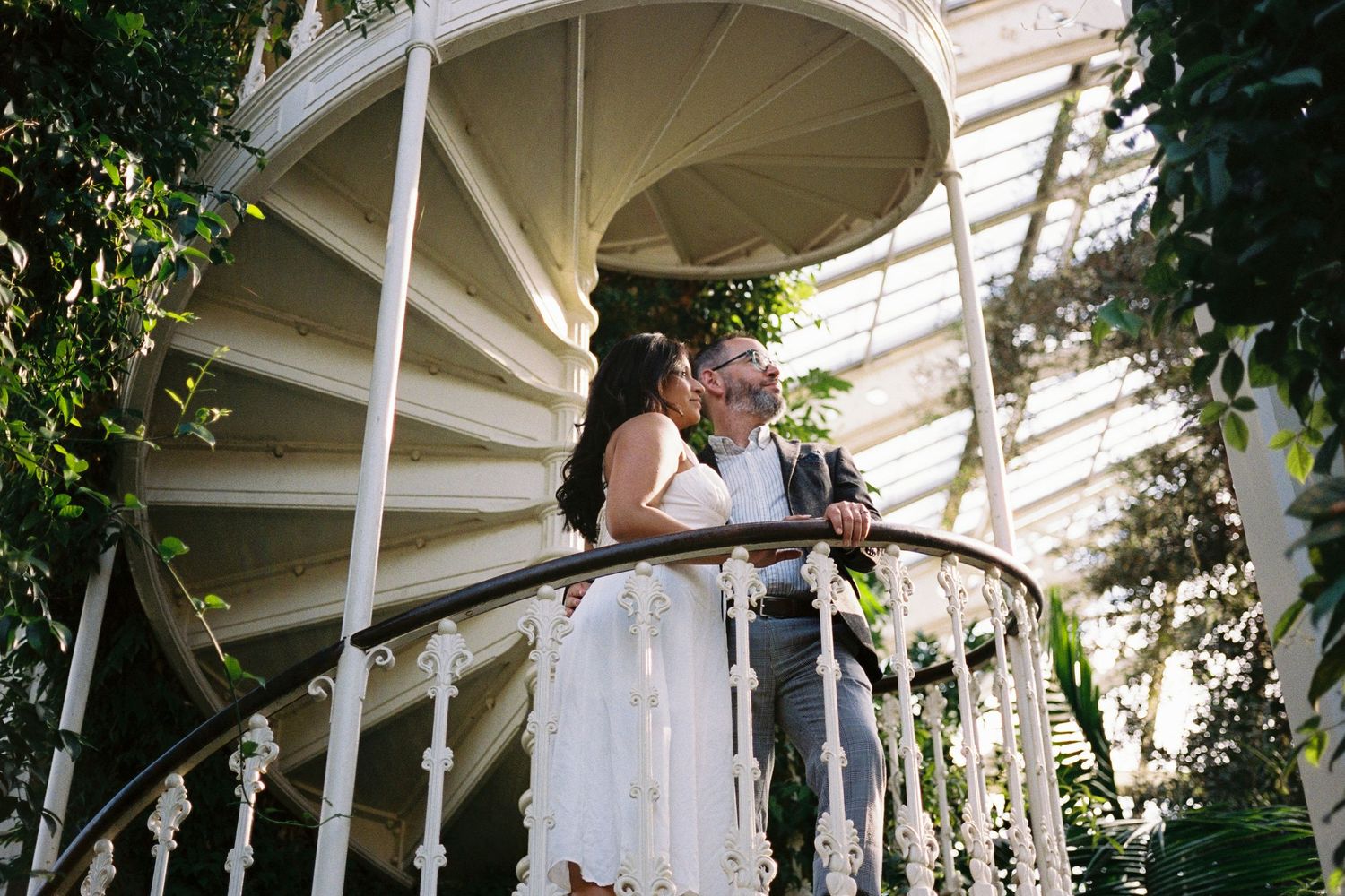 Bride and groom share a romantic kiss on a white spiral staircase surrounded by lush greenery.
