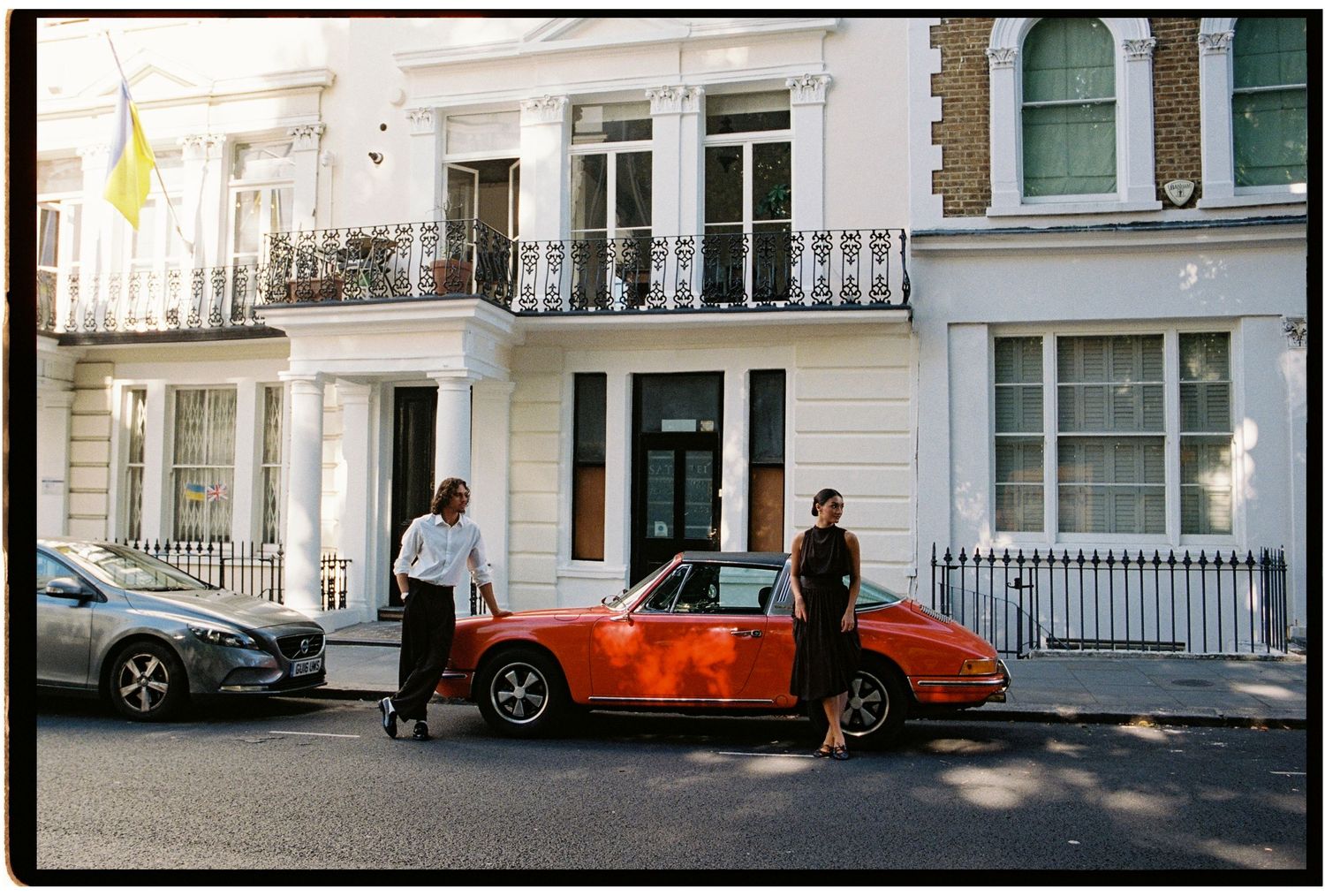 Two people standing beside a vintage red Porsche on a London street with white Georgian townhouses in the background.