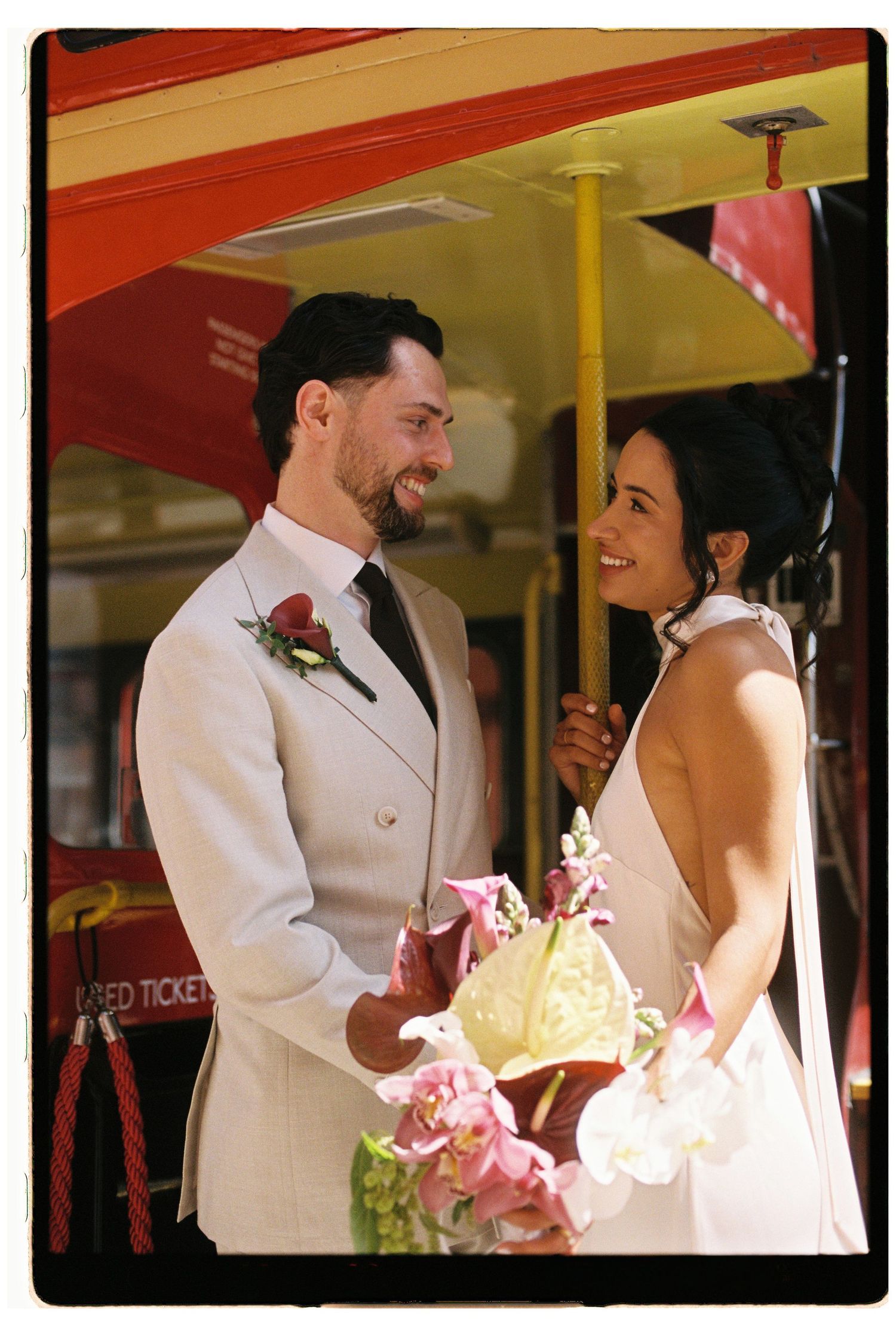 Bride and groom sharing a romantic moment beside a vintage red London bus, bride holding tropical floral bouquet.
