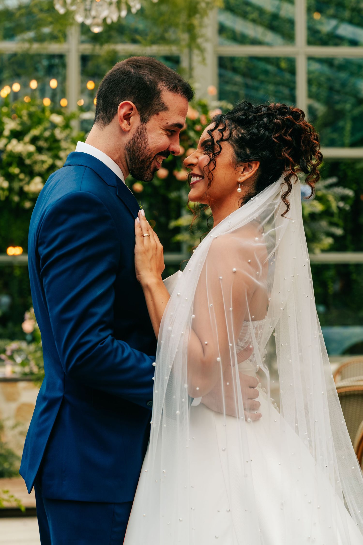 Smiling bride in pearl veil and groom in navy suit share tender moment in garden greenhouse.