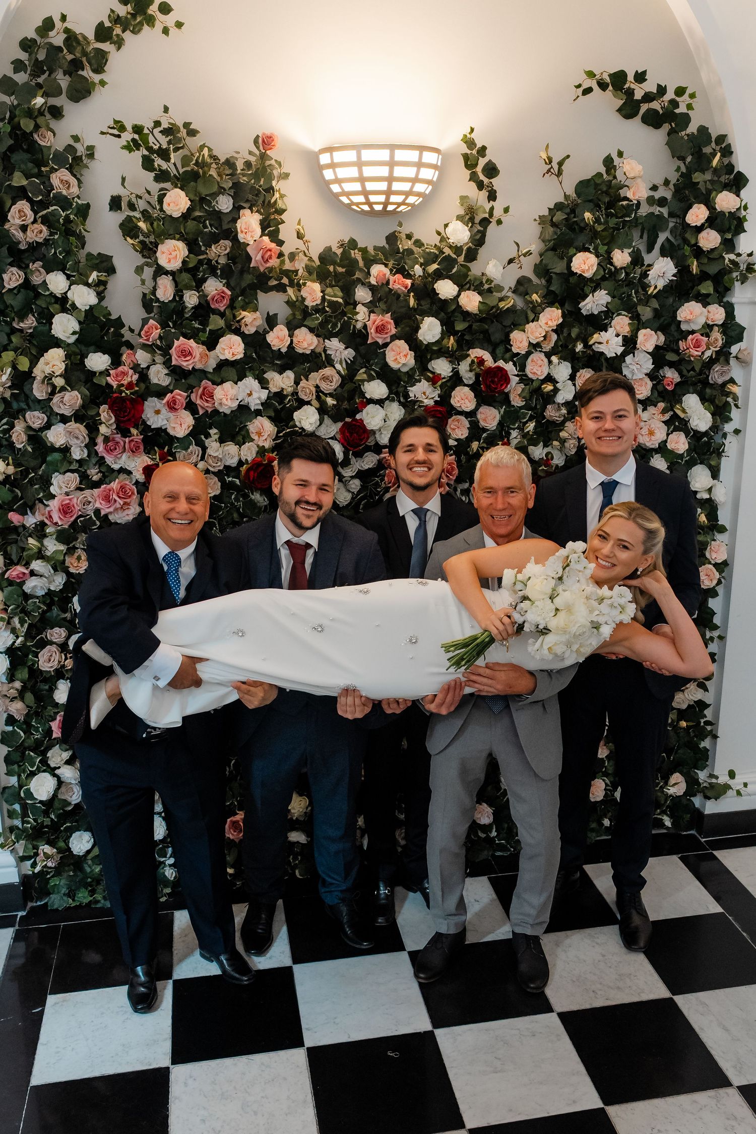Groom and groomsmen playfully carrying the bride in front of a floral wall backdrop.
