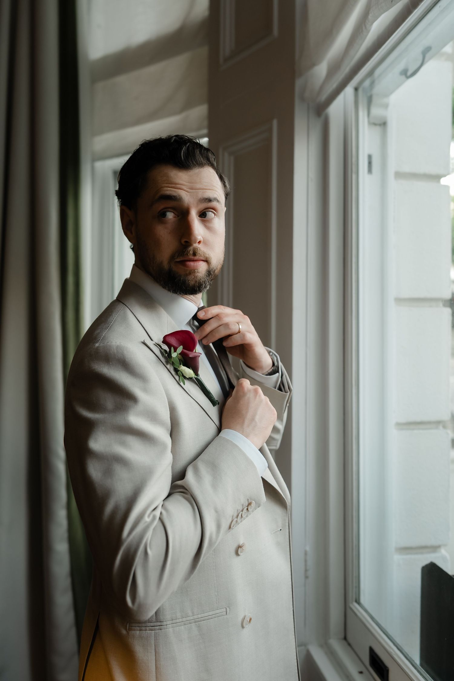 Handsome bearded groom in a beige suit adjusting his tie while gazing out a bright window on wedding day.