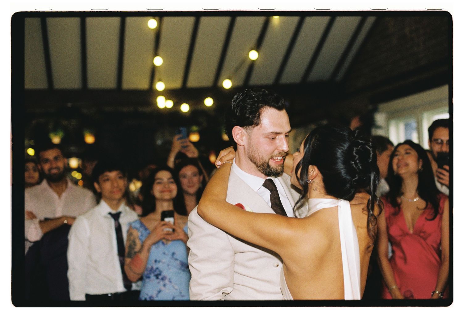 Couple sharing first dance embracing closely surrounded by guests at wedding reception.