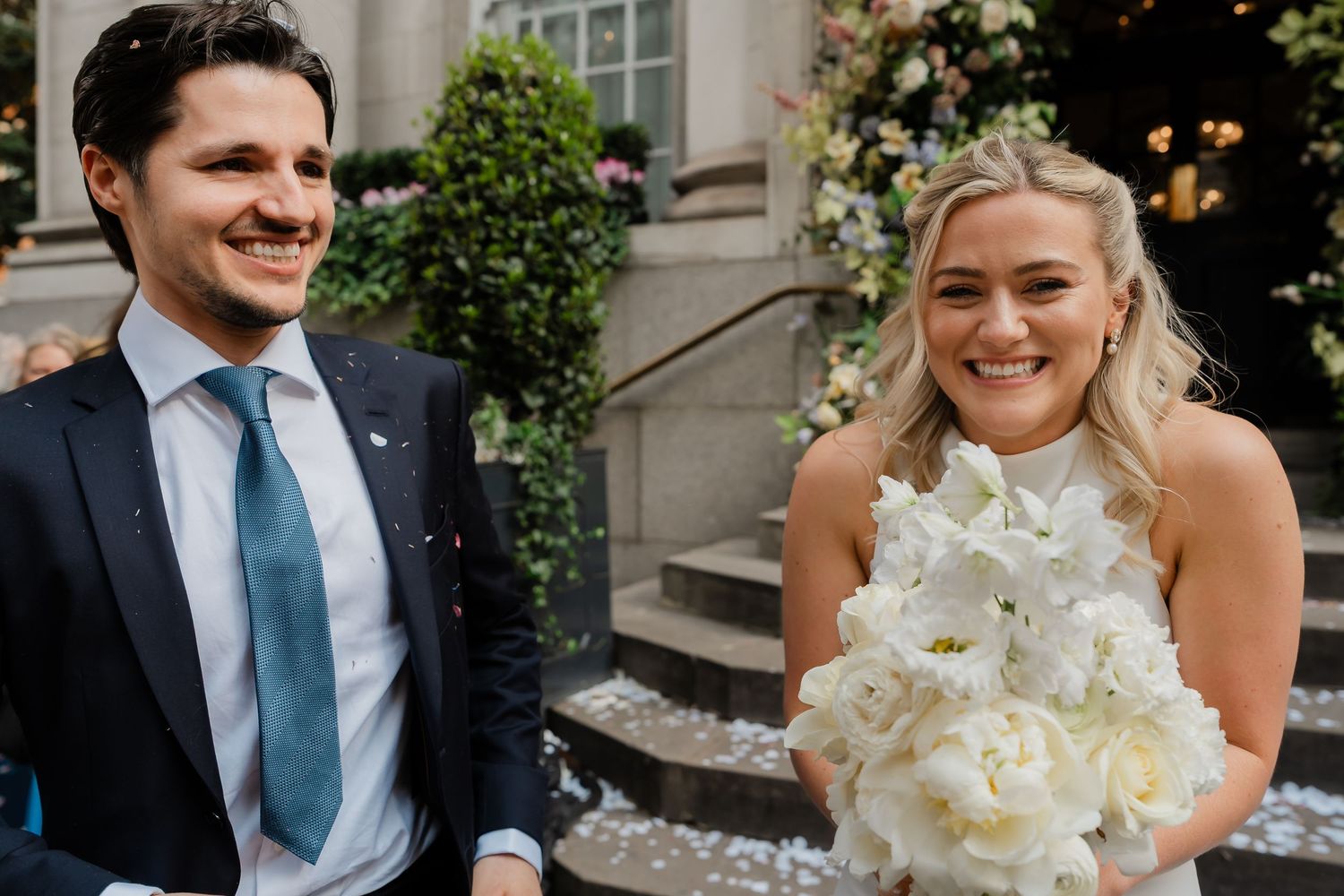 Smiling bride holding white bouquet stands beside groom in navy suit after ceremony.