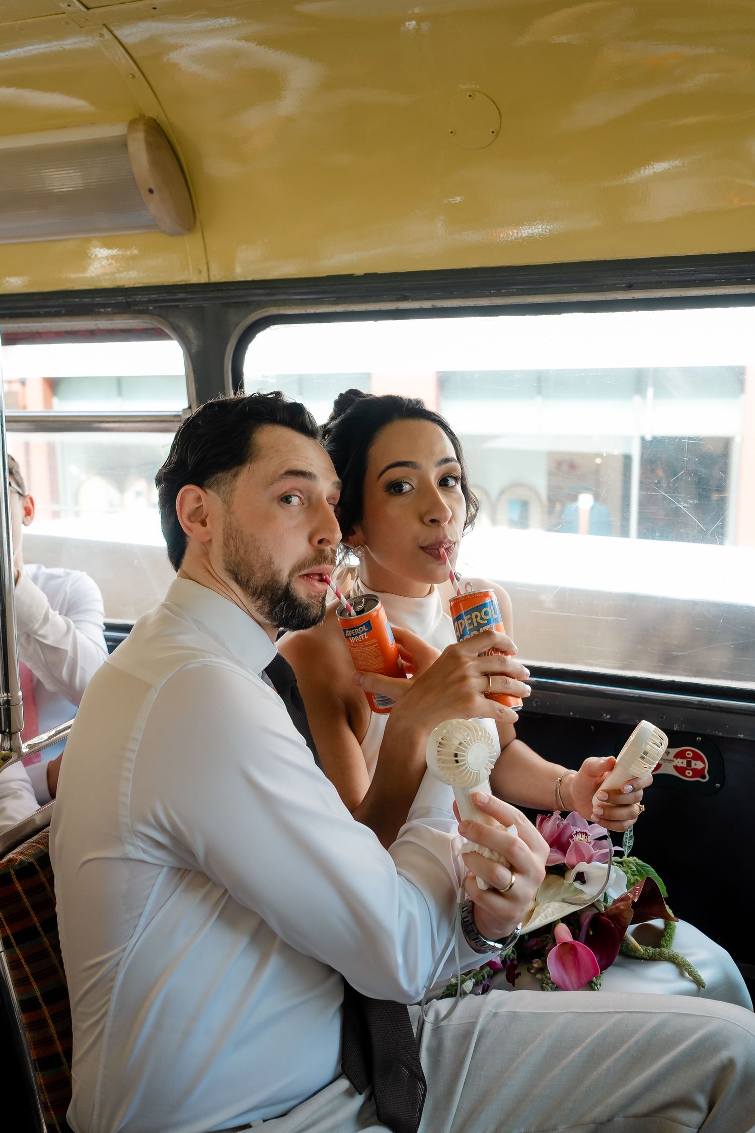 Bride and groom sipping drinks through straws together while seated inside a vintage double-decker wedding bus.