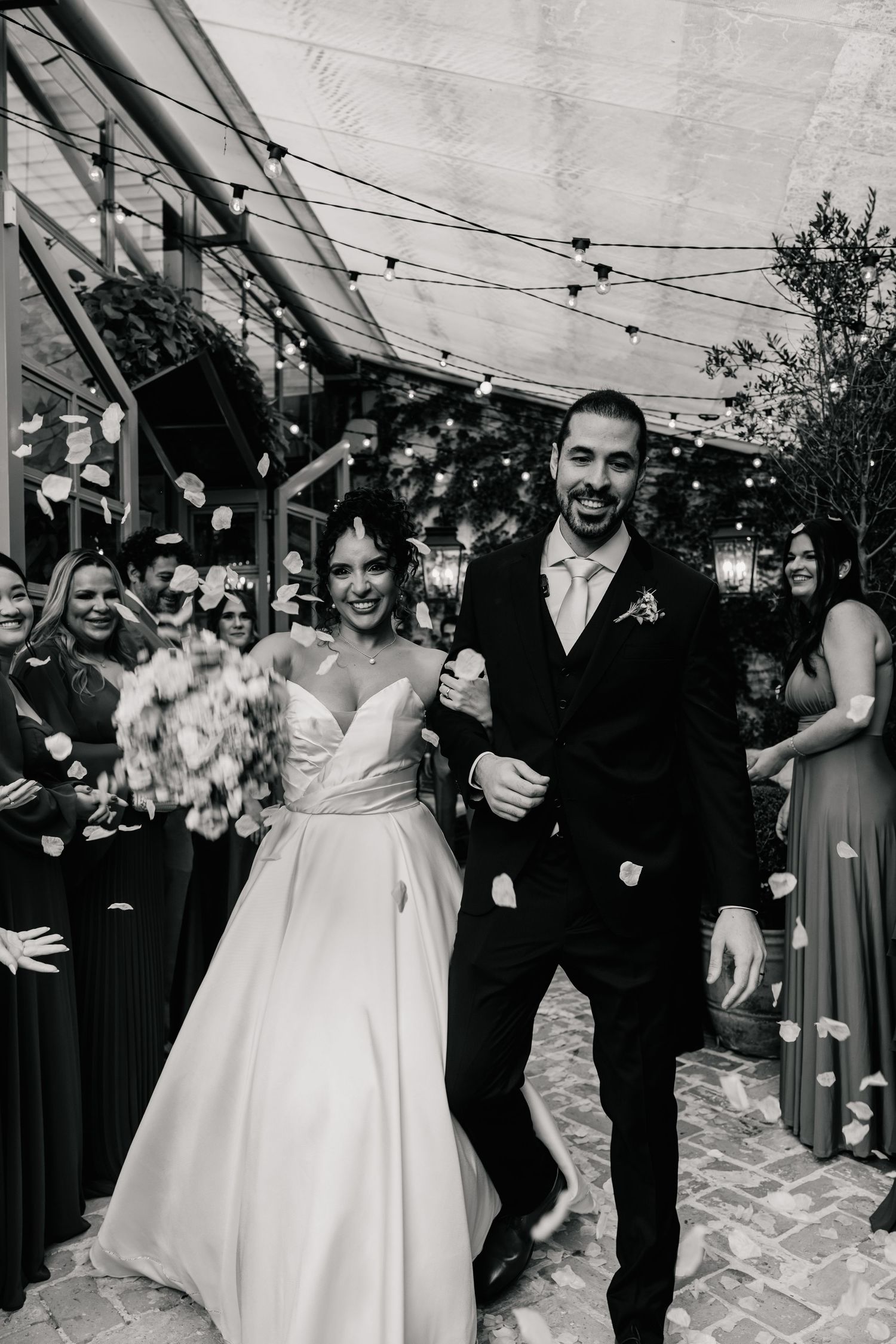 Black and white photo of smiling bride and groom walking through confetti toss at wedding reception.
