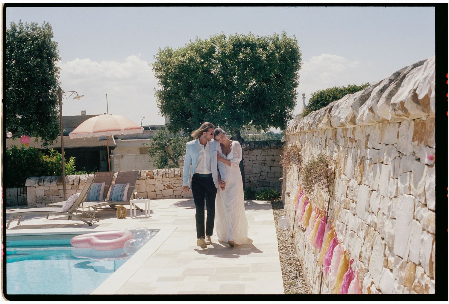 Couple sharing a romantic moment beside a sunlit pool with stone walls and colorful decorations at an outdoor wedding venue.