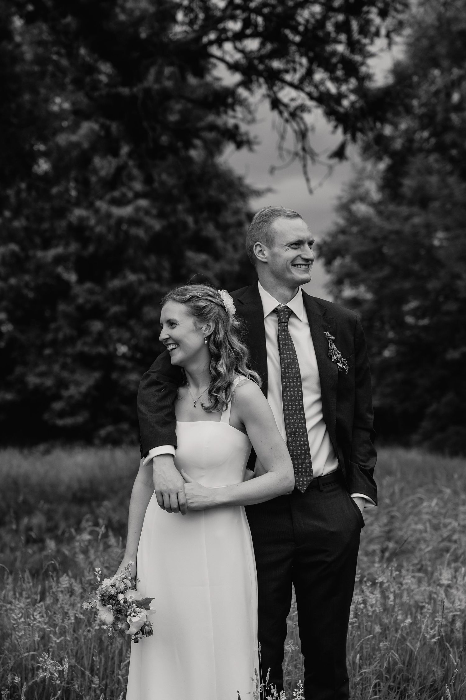 A black and white photo of a smiling bride and groom embracing in a meadow surrounded by tall trees.