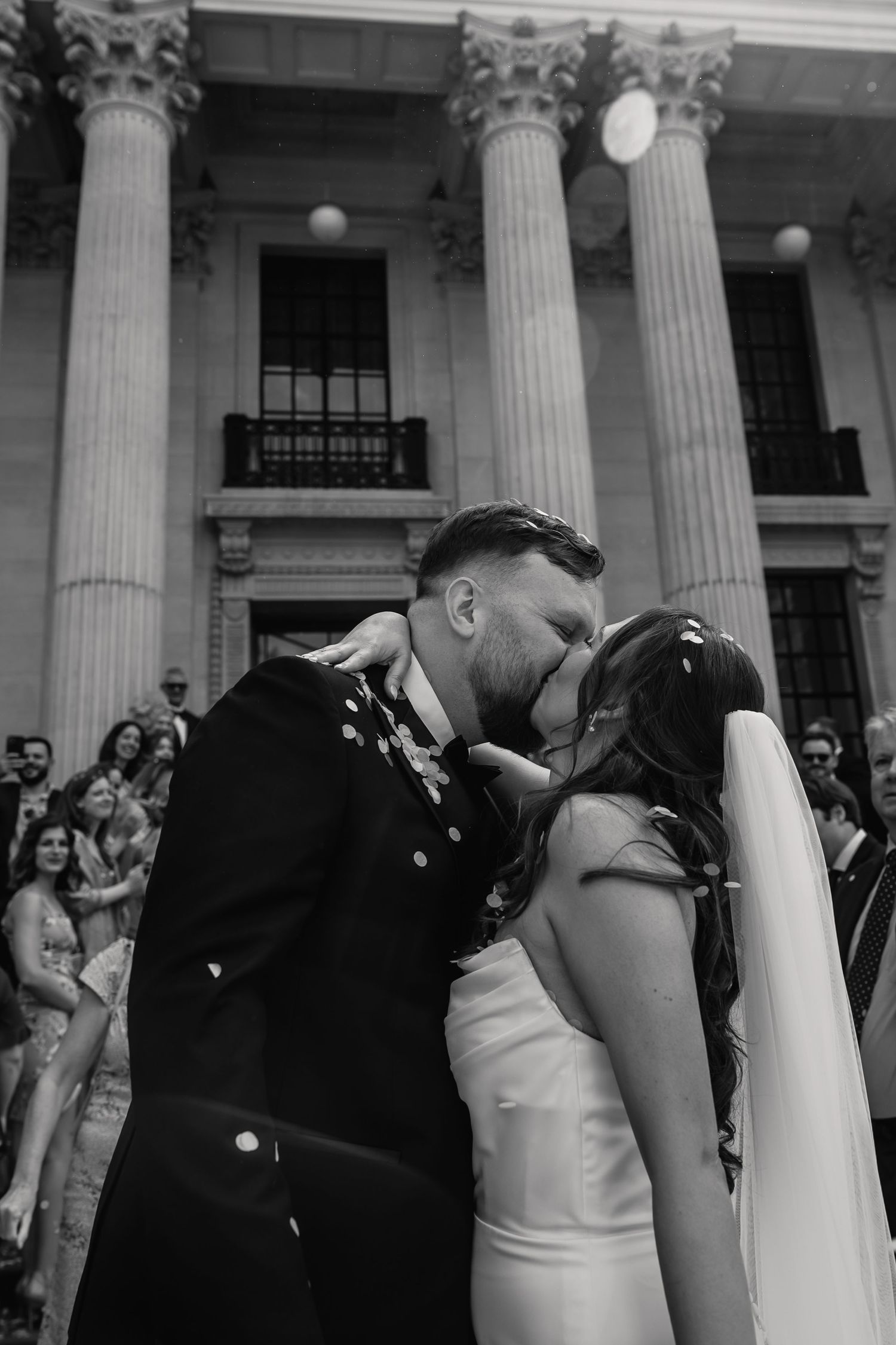 Black and white photo of bride and groom sharing first kiss outside neoclassical wedding venue.
