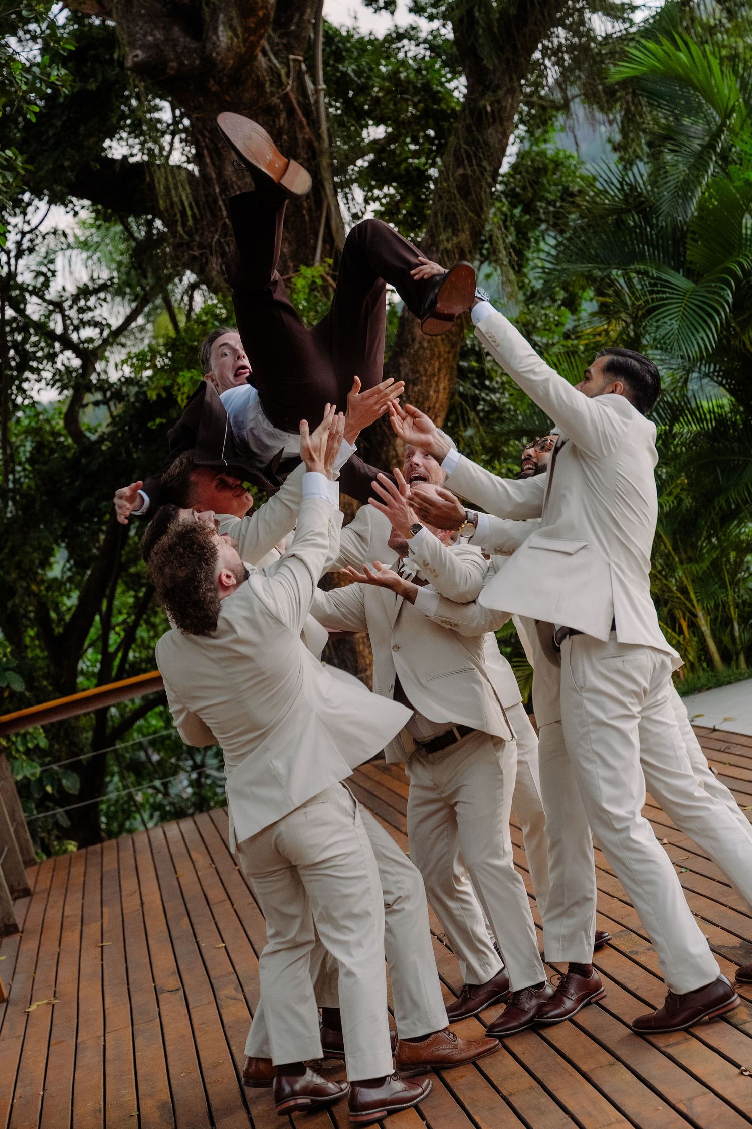 Groomsmen in white suits toss groom into air on wooden deck surrounded by tropical greenery.