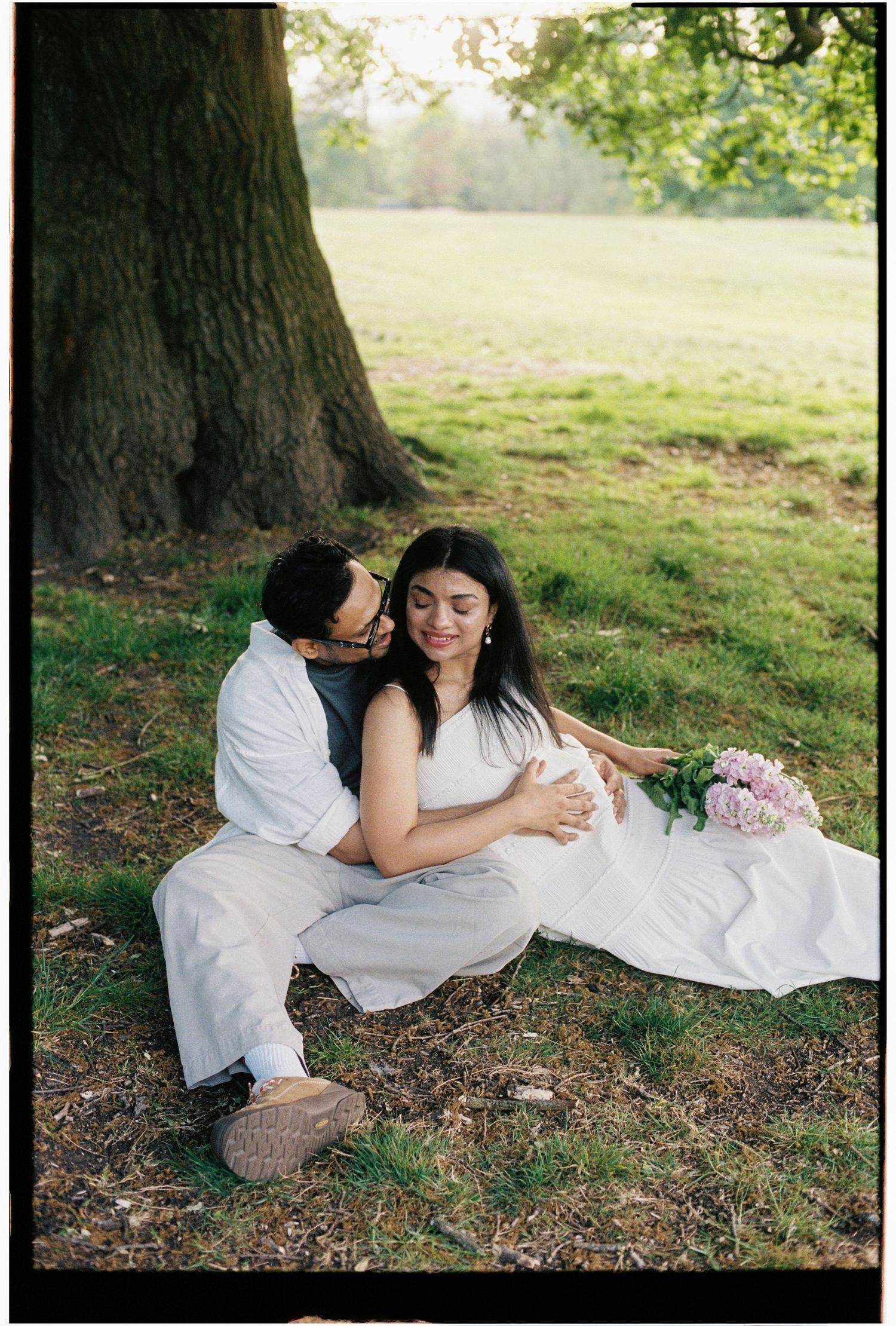 Couple sitting under large tree in park, man embracing pregnant woman with pink peonies nearby.