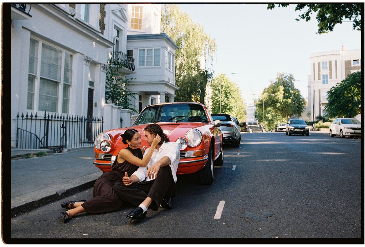 Couple kissing and embracing in front of a vintage red Porsche on a quiet tree-lined London street.