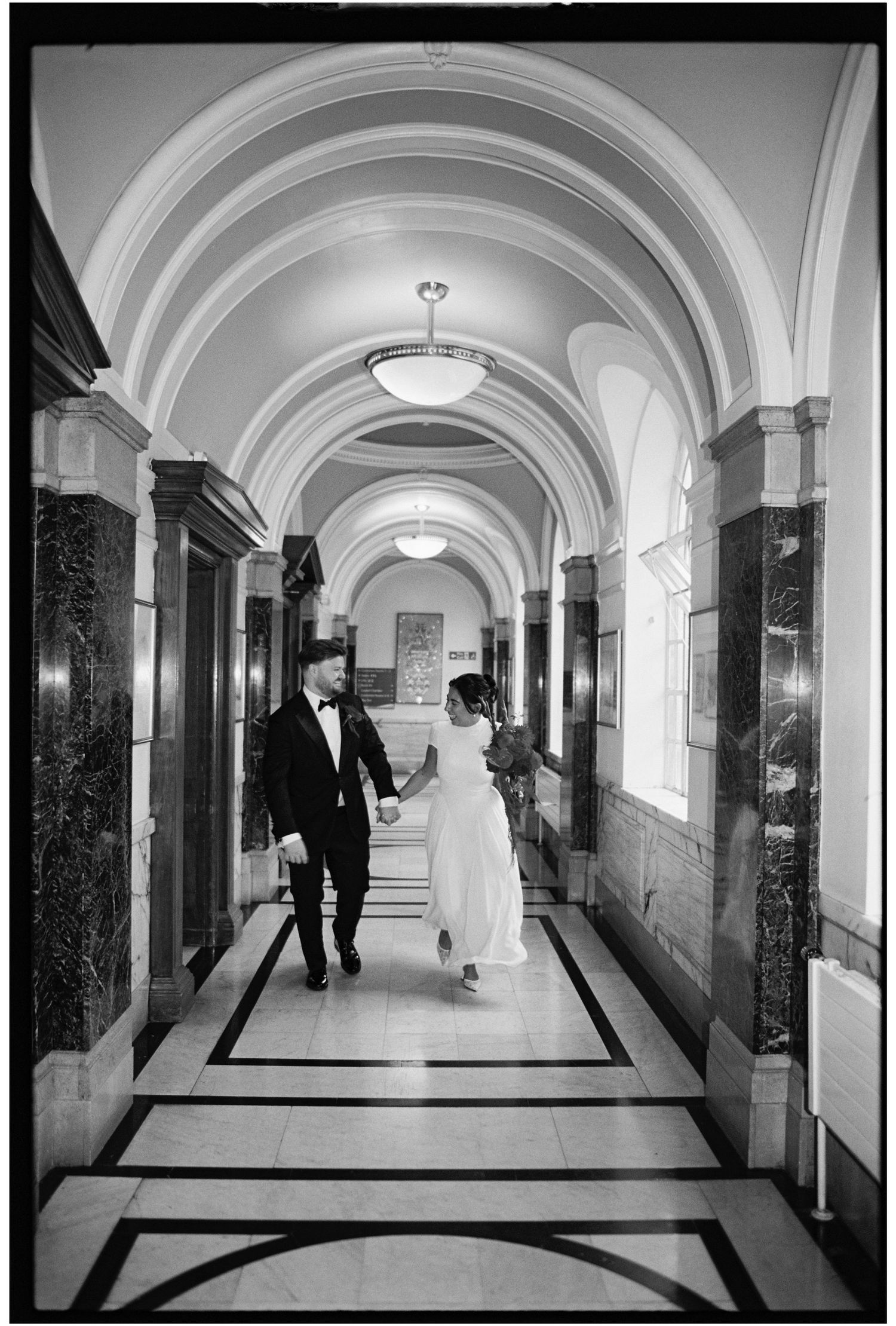 Black and white photo of a bride and groom holding hands walking through an elegant arched hallway.