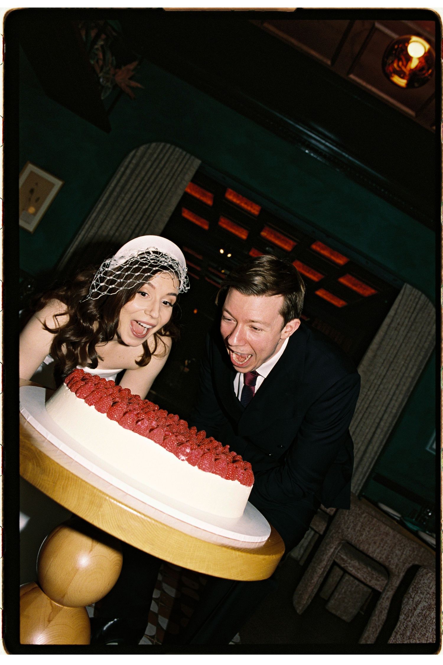 Bride and groom laughing excitedly over a white wedding cake decorated with red roses at their reception.