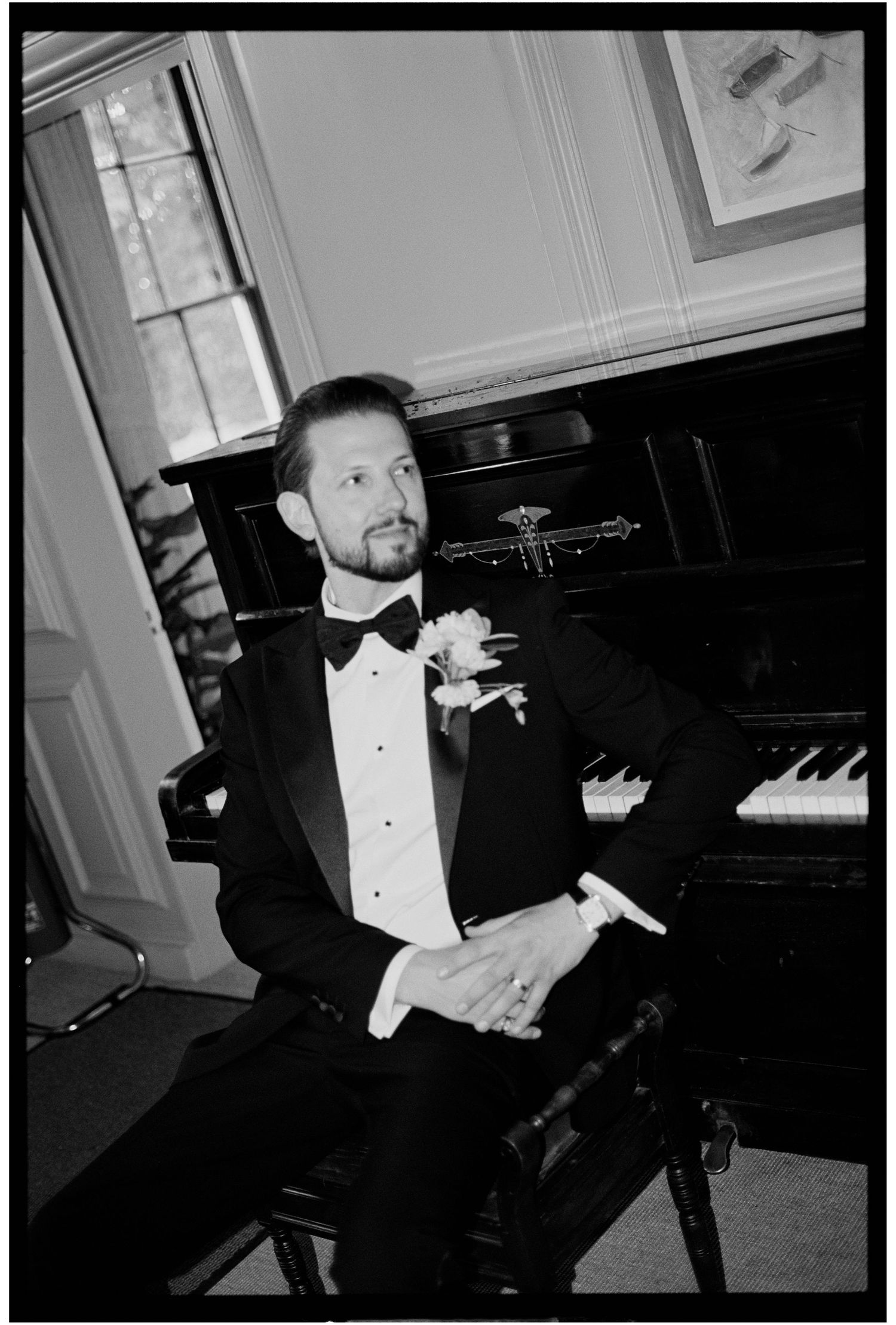 Elegant groom in black tuxedo with bow tie and boutonniere seated beside a grand piano in black and white photo.