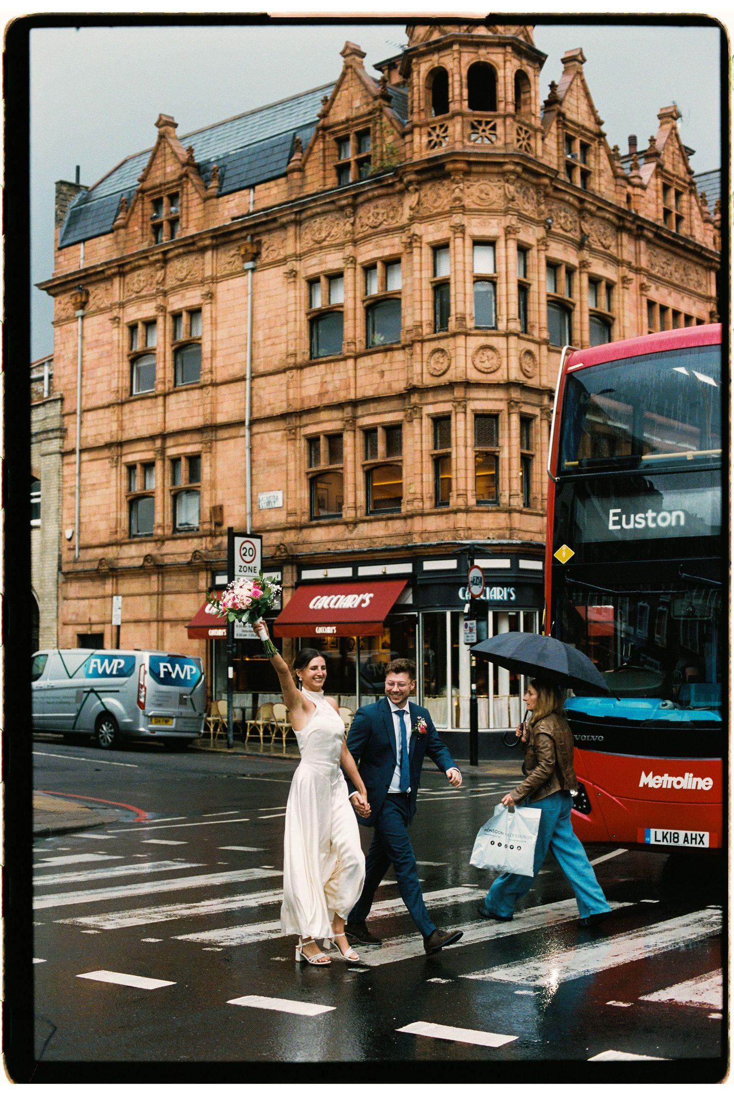 Bride and groom crossing a zebra crossing in London with a red double-decker Euston bus and Victorian building behind them.