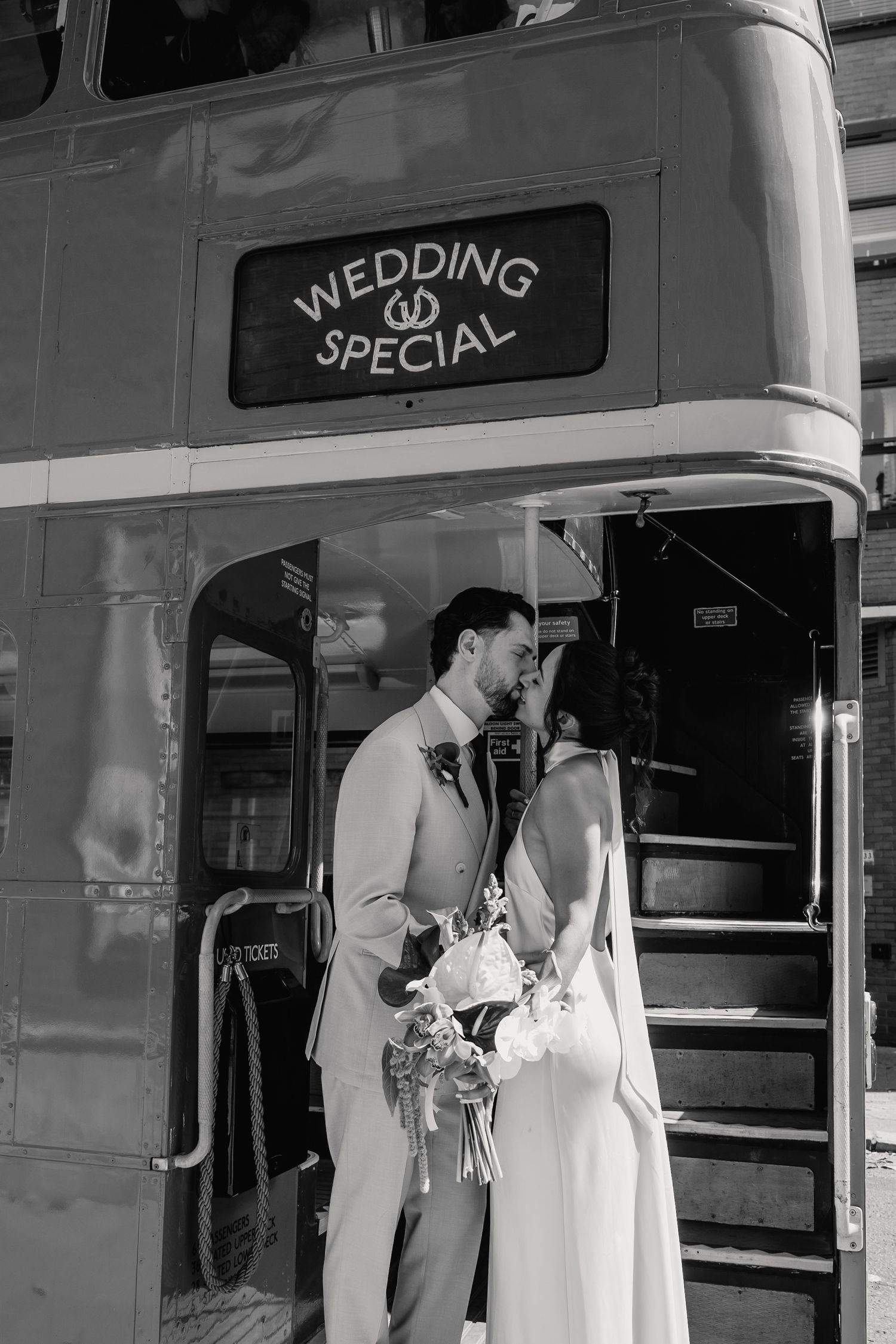 Black and white photo of bride and groom sharing a kiss at the entrance of a wedding special double-decker bus.