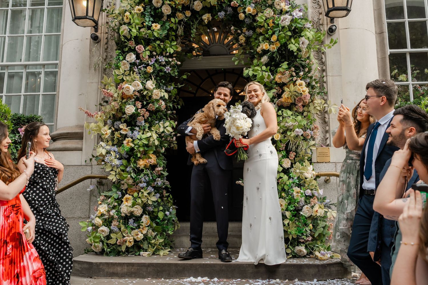 Bride and groom embrace with small dog outside beautifully flower-decorated venue entrance.