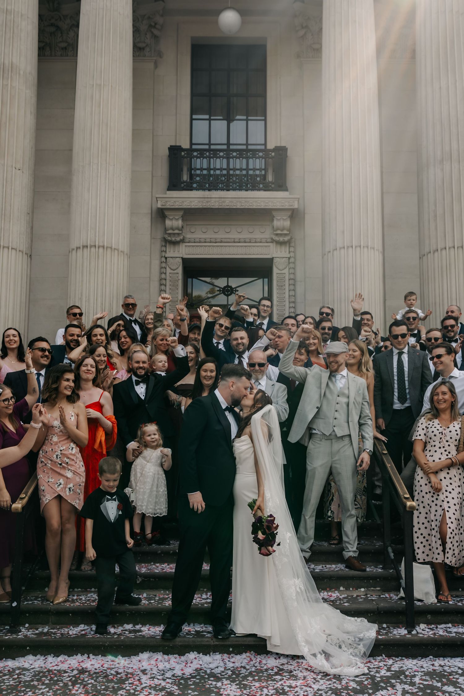 Newlyweds kiss on steps surrounded by cheering wedding guests throwing confetti at elegant venue.