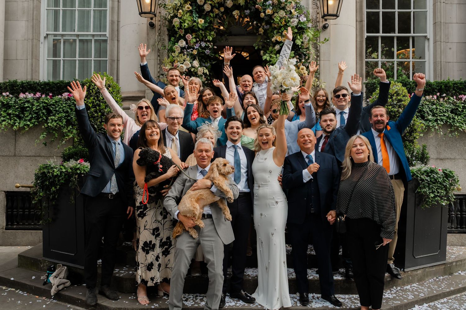 Wedding party group photo with guests cheering outside a elegant venue decorated with flowers.