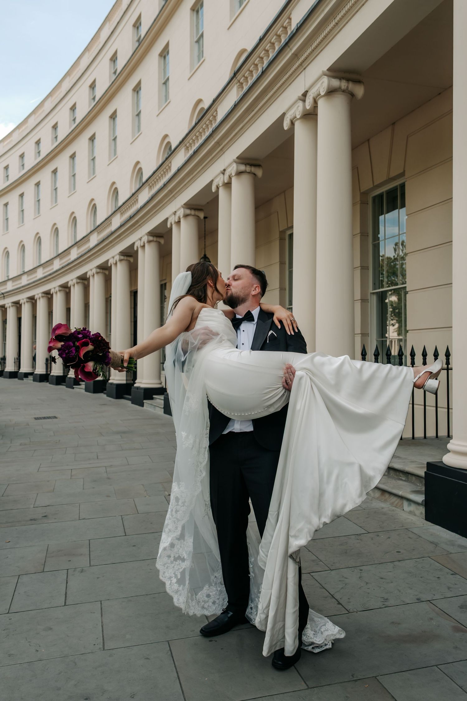 Groom lifts bride in wedding dress outside grand curved neoclassical building with tall white columns.