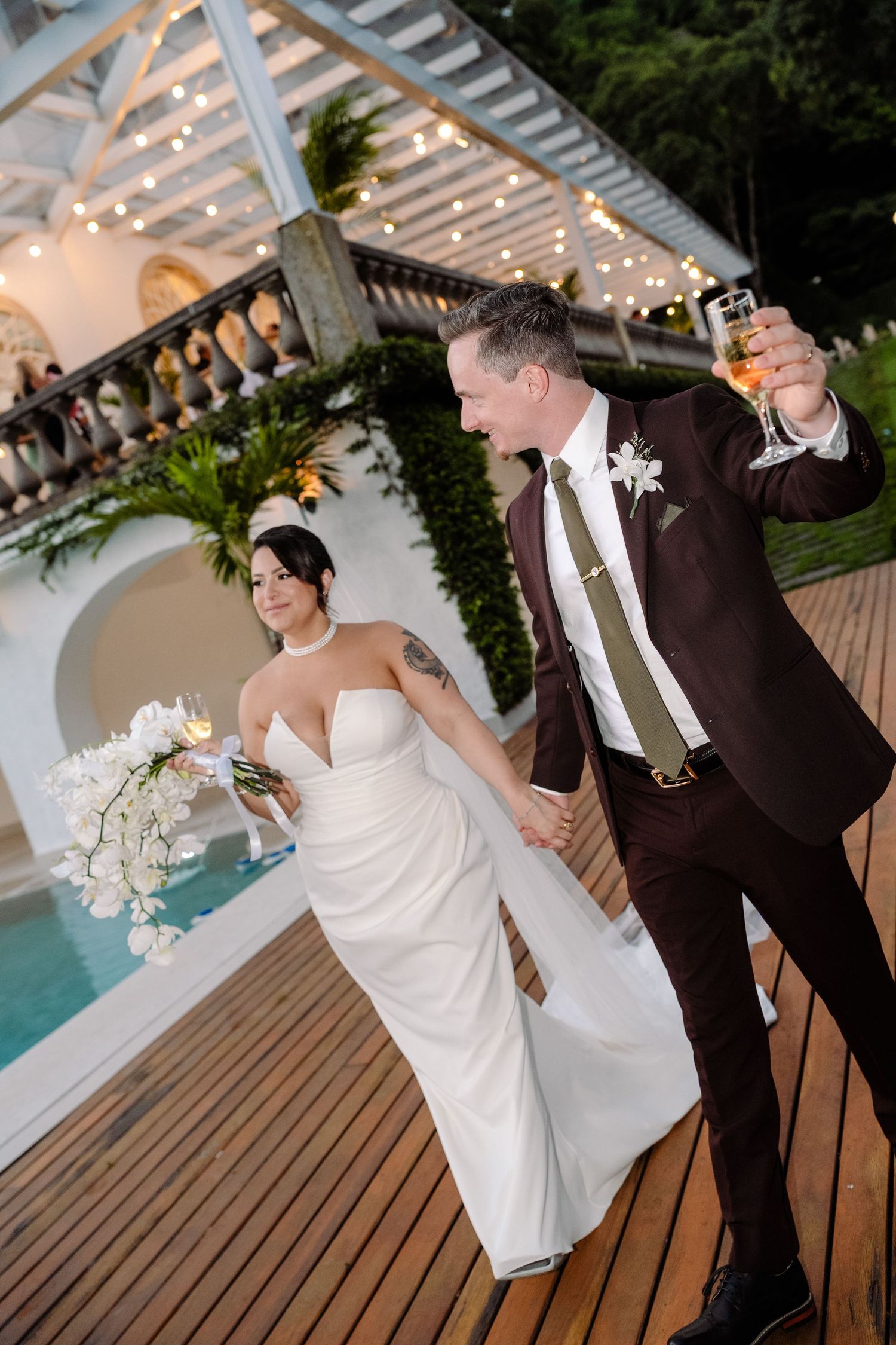 Bride and groom celebrate by poolside at night, groom raising champagne glass at elegant venue.
