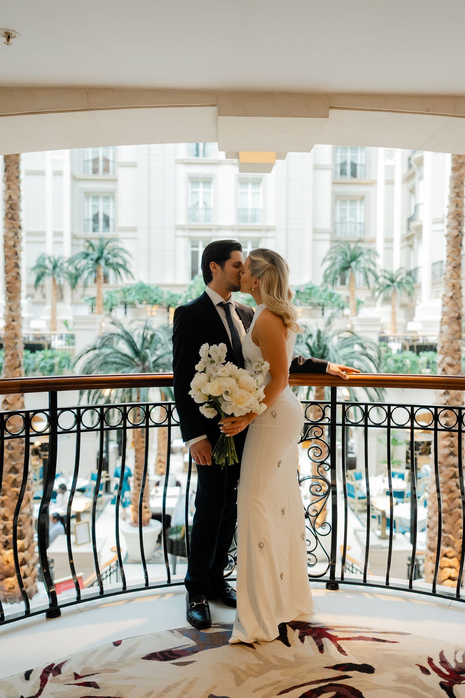 Bride and groom share a romantic kiss on an elegant balcony with palm trees and a grand building backdrop.