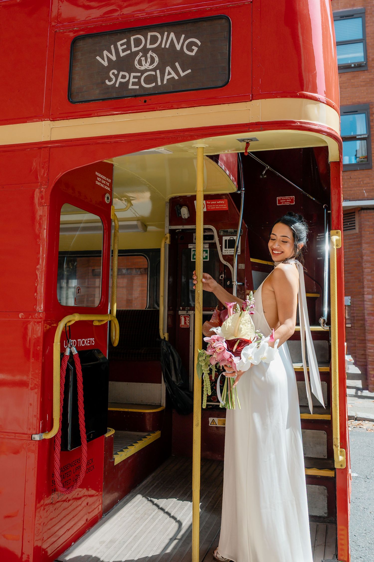 Smiling bride in backless white gown boarding a red London double-decker wedding special bus holding floral bouquet.