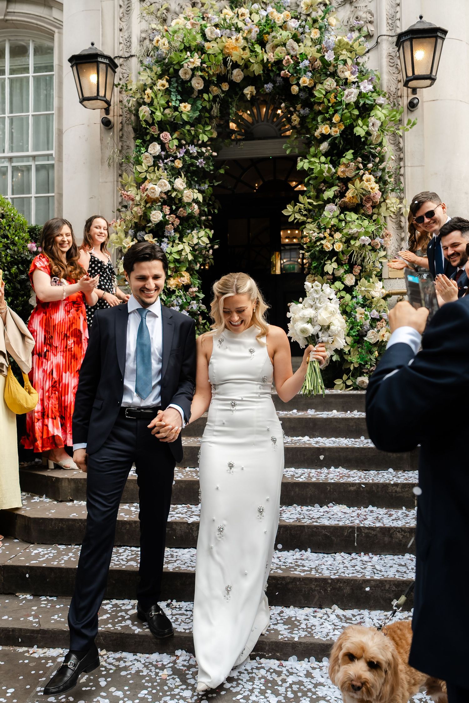 Newlyweds descend flower-covered venue steps hand in hand amid confetti celebration.
