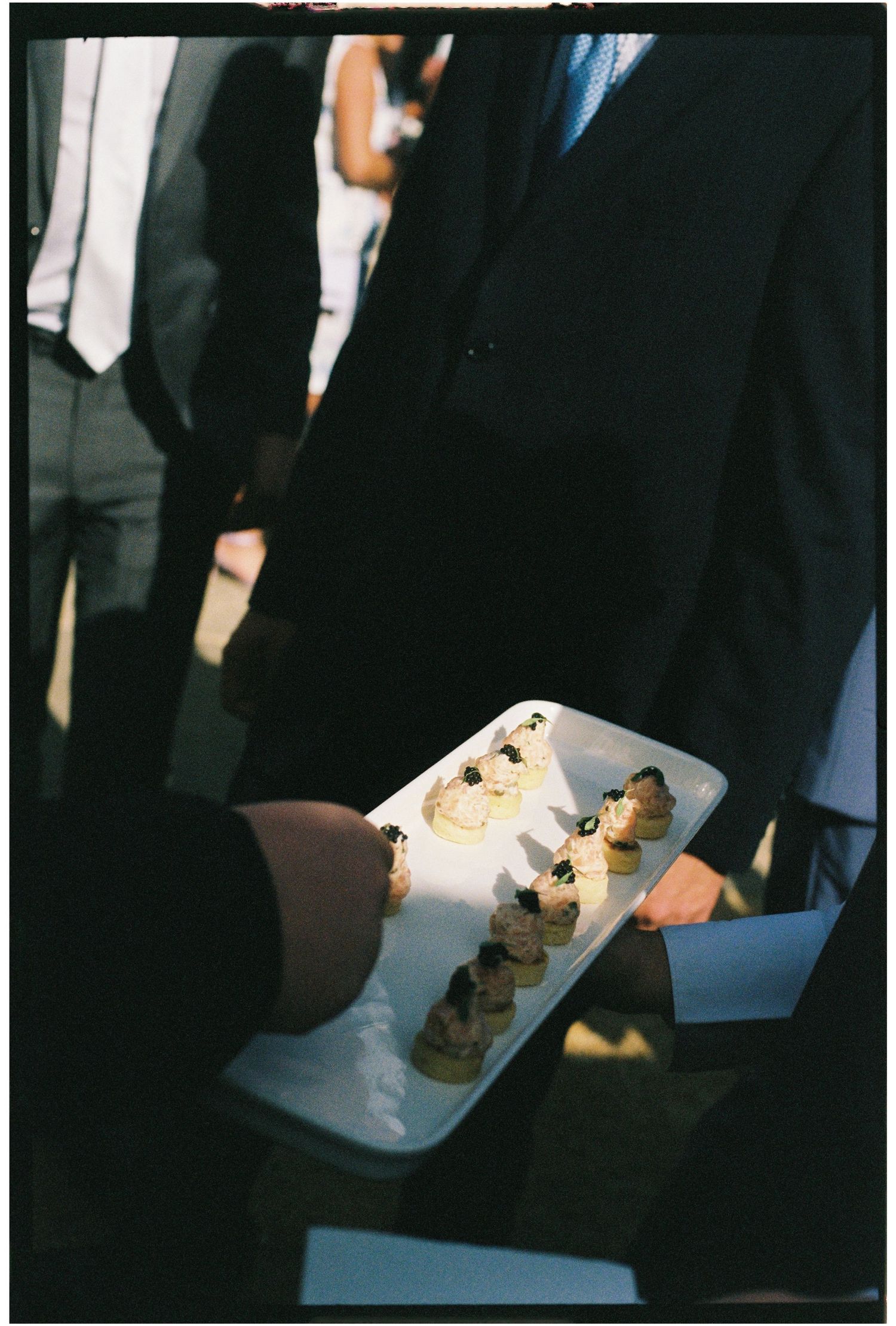 A server holds a white tray of elegant canapés at a formal event, surrounded by guests in suits.