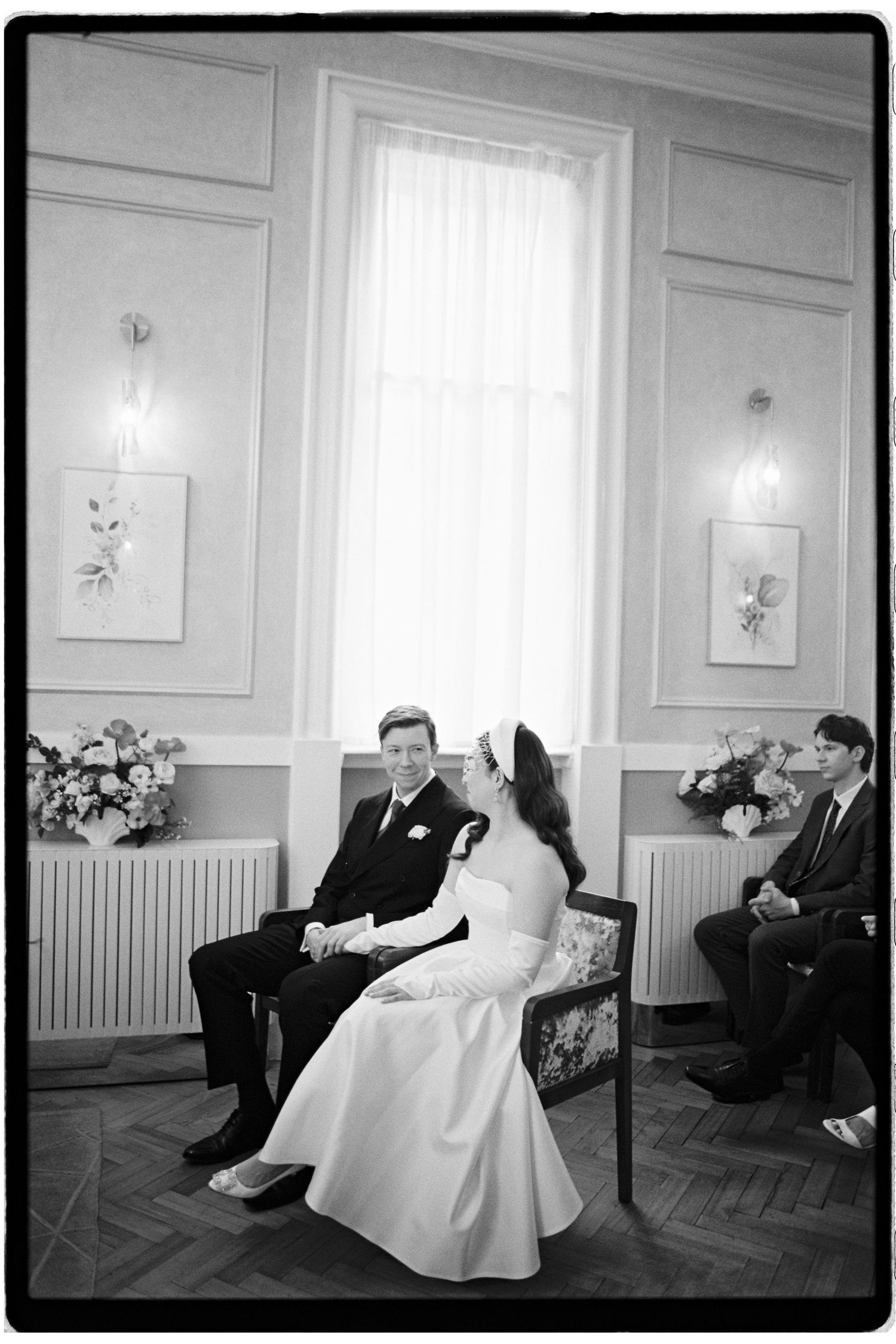 Black and white photo of a bride and groom sitting together at a wedding ceremony in an elegant room.