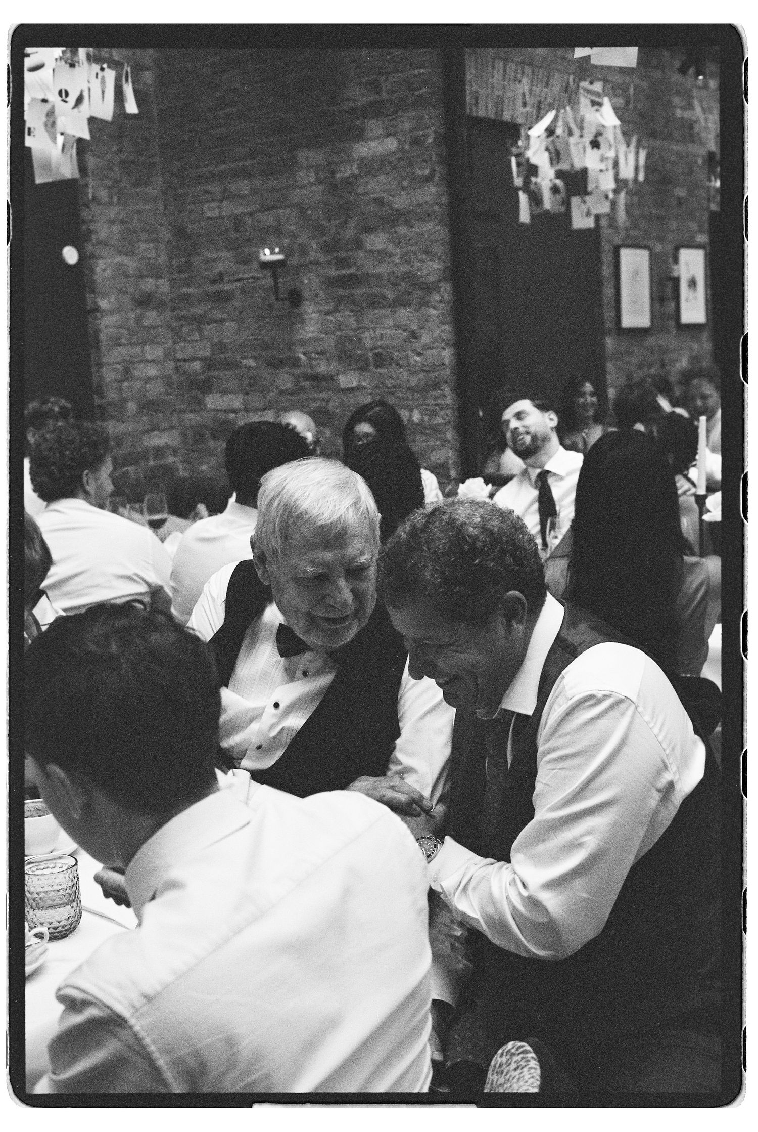 Black and white candid photo of elderly wedding guests in conversation at rustic brick-walled reception venue.