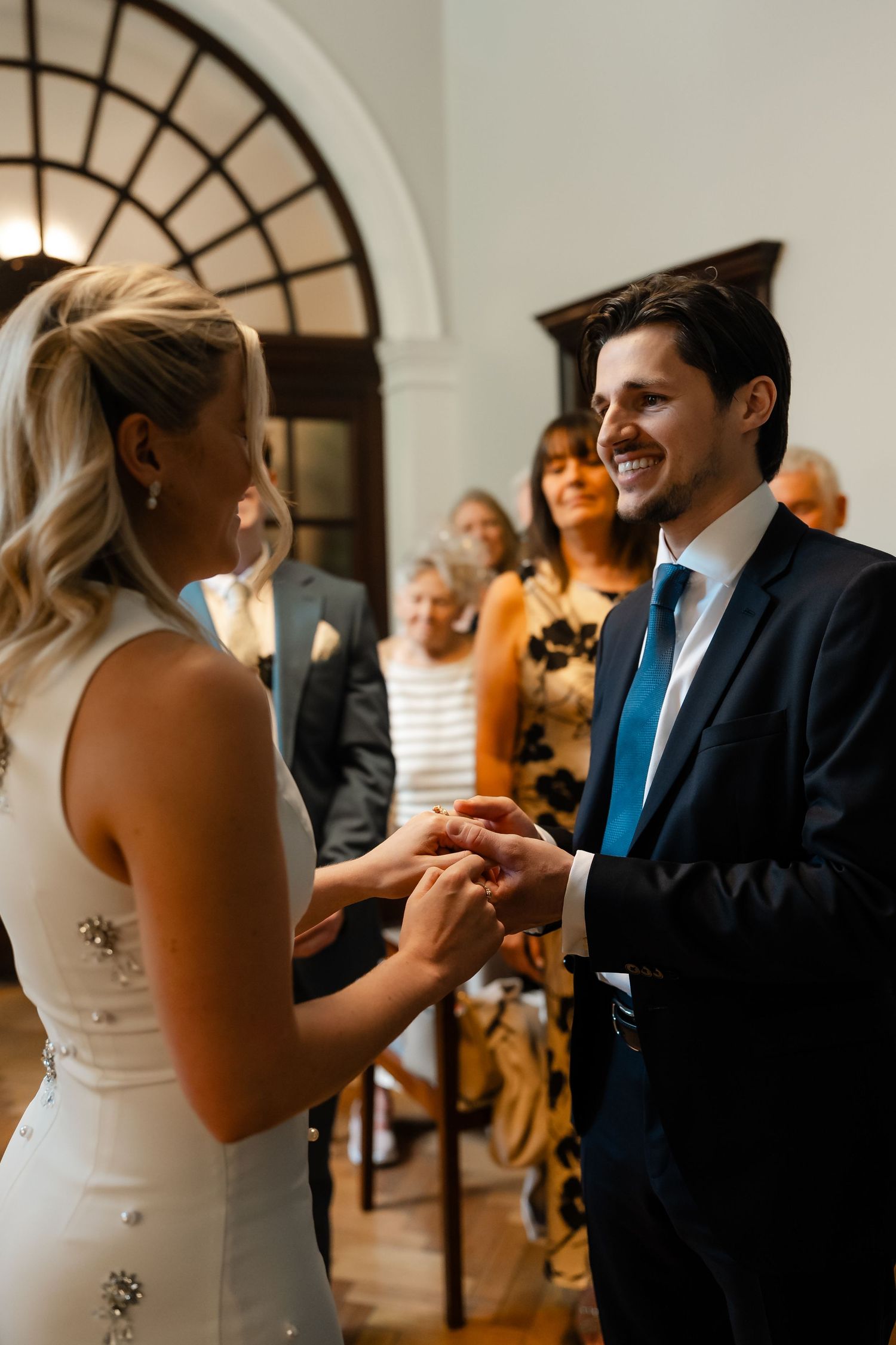 Groom smiling as bride places wedding ring on his finger during indoor ceremony.