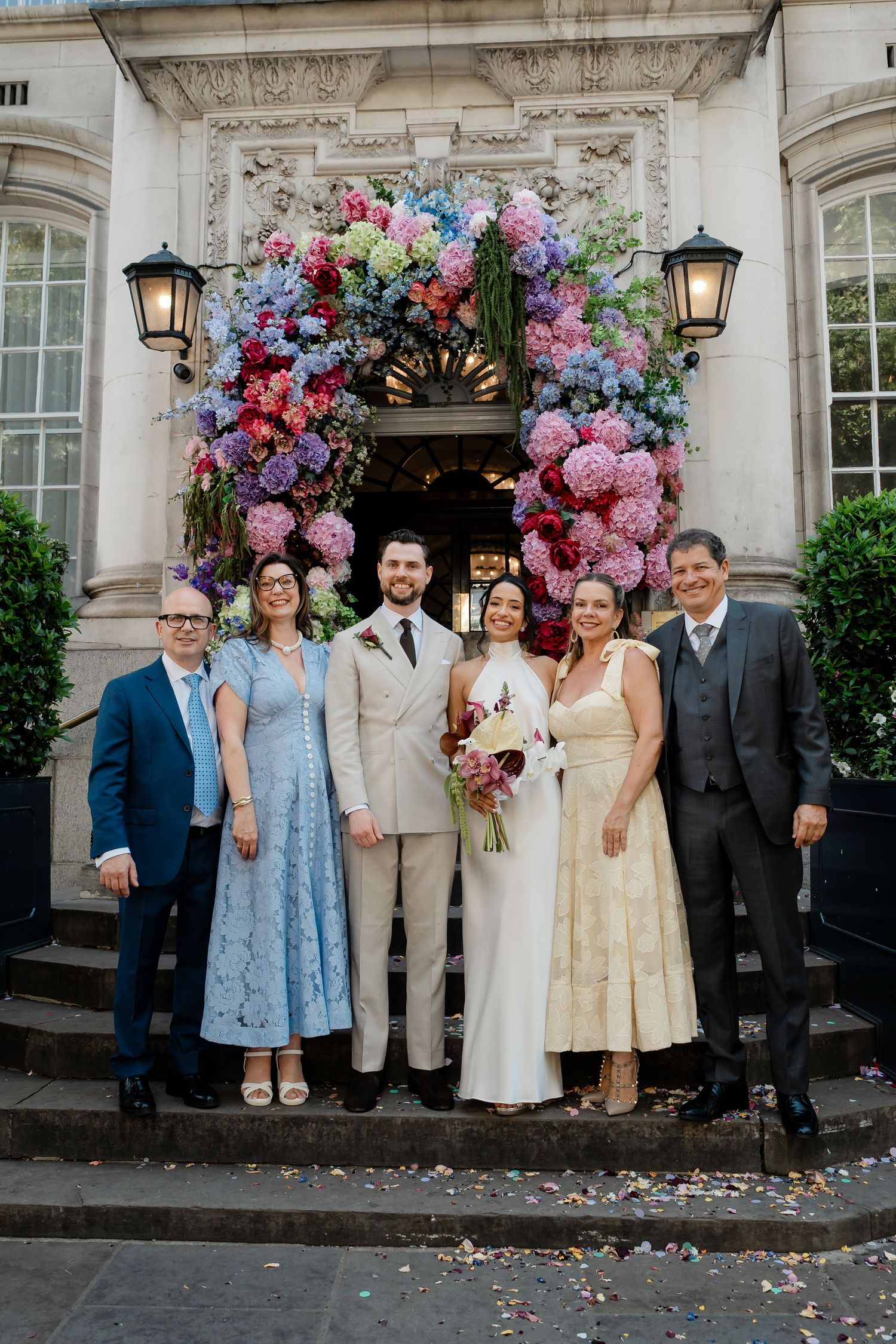 Wedding party posing before colorful floral arch at elegant venue entrance.