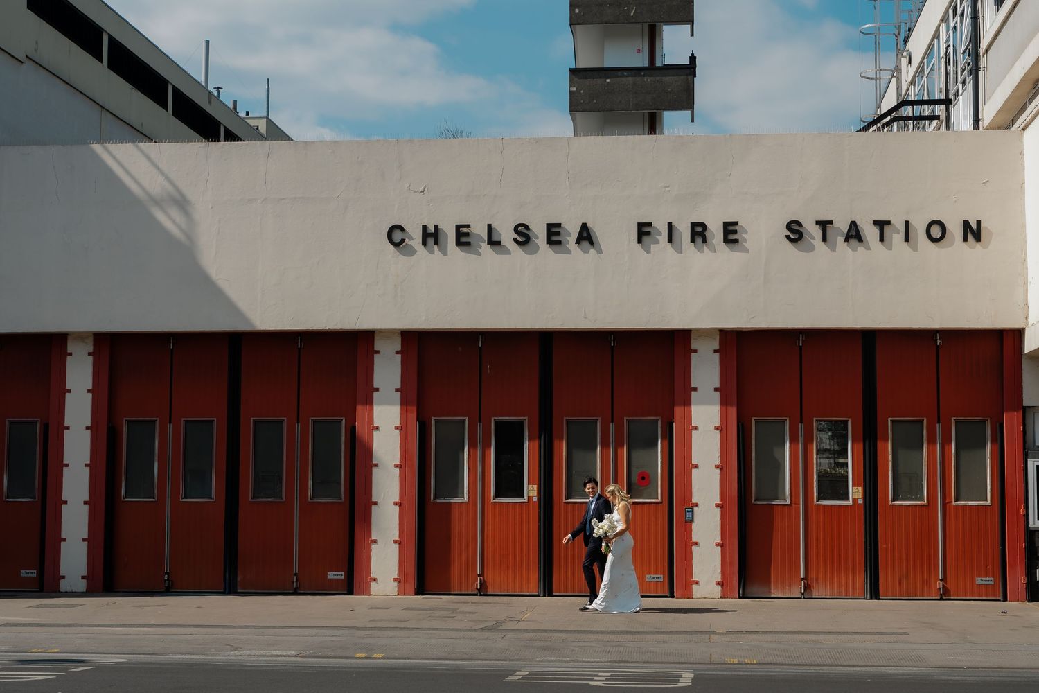 Bride and groom embrace outside Chelsea Fire Station with iconic red garage doors in London.