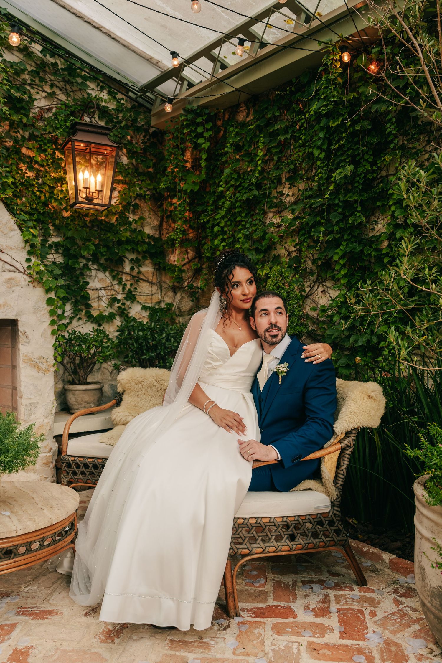 Bride and groom pose together on wicker chair surrounded by lush ivy-covered walls.