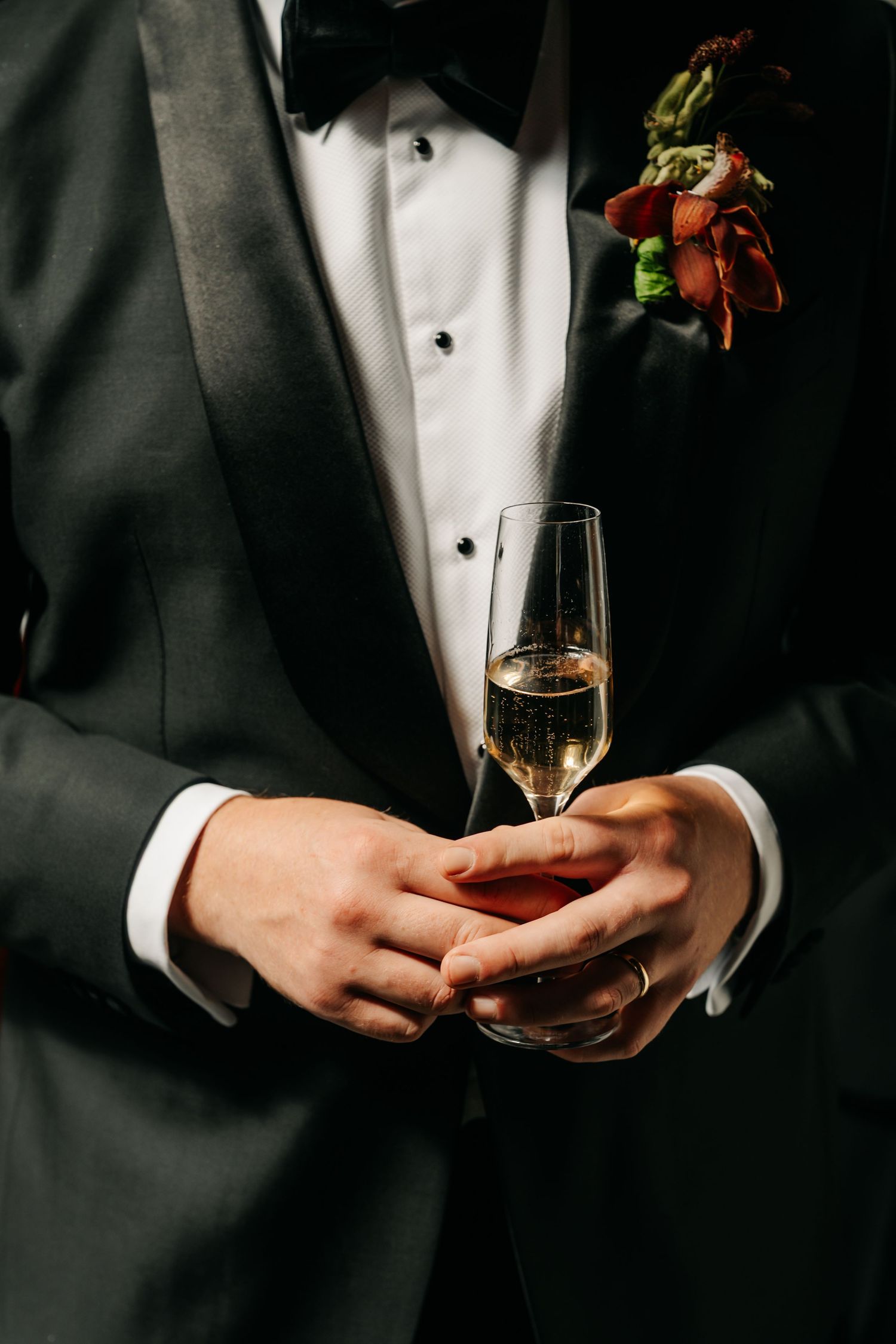Groom in black tuxedo holds champagne flute with floral boutonniere at wedding.