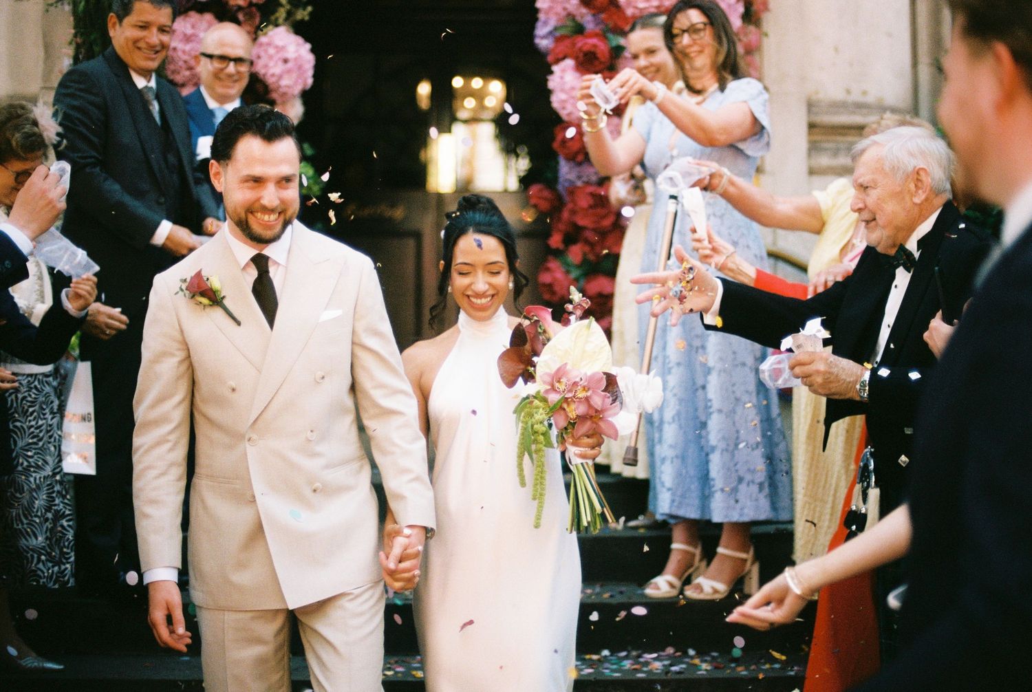 Newlyweds walk hand in hand through a shower of confetti as guests celebrate outside a floral-adorned venue.