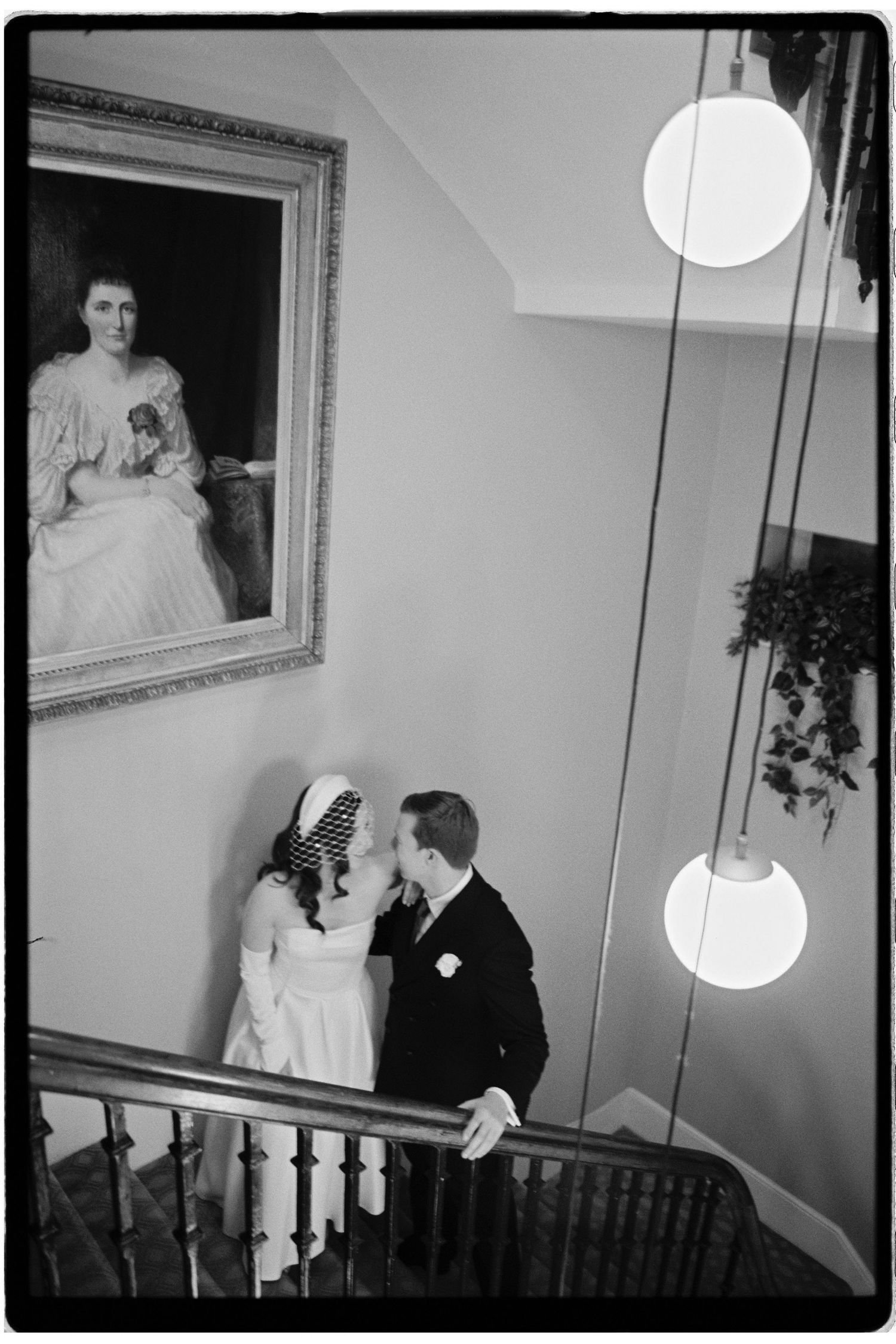Black and white photo of a bride and groom sharing a moment on a staircase, with a portrait painting on the wall above.