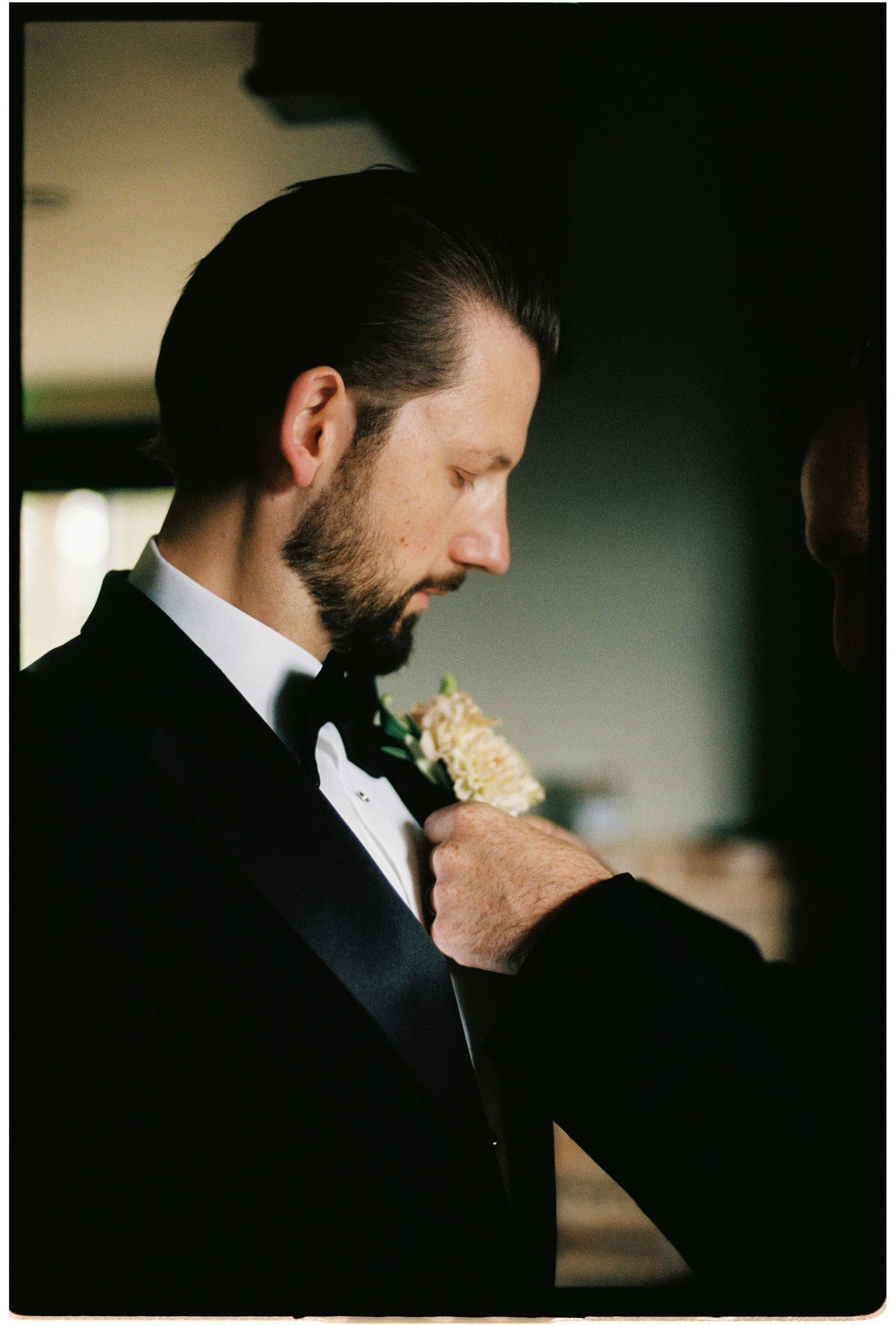 Groom in black tuxedo adjusts his floral boutonniere while looking down in soft natural light.
