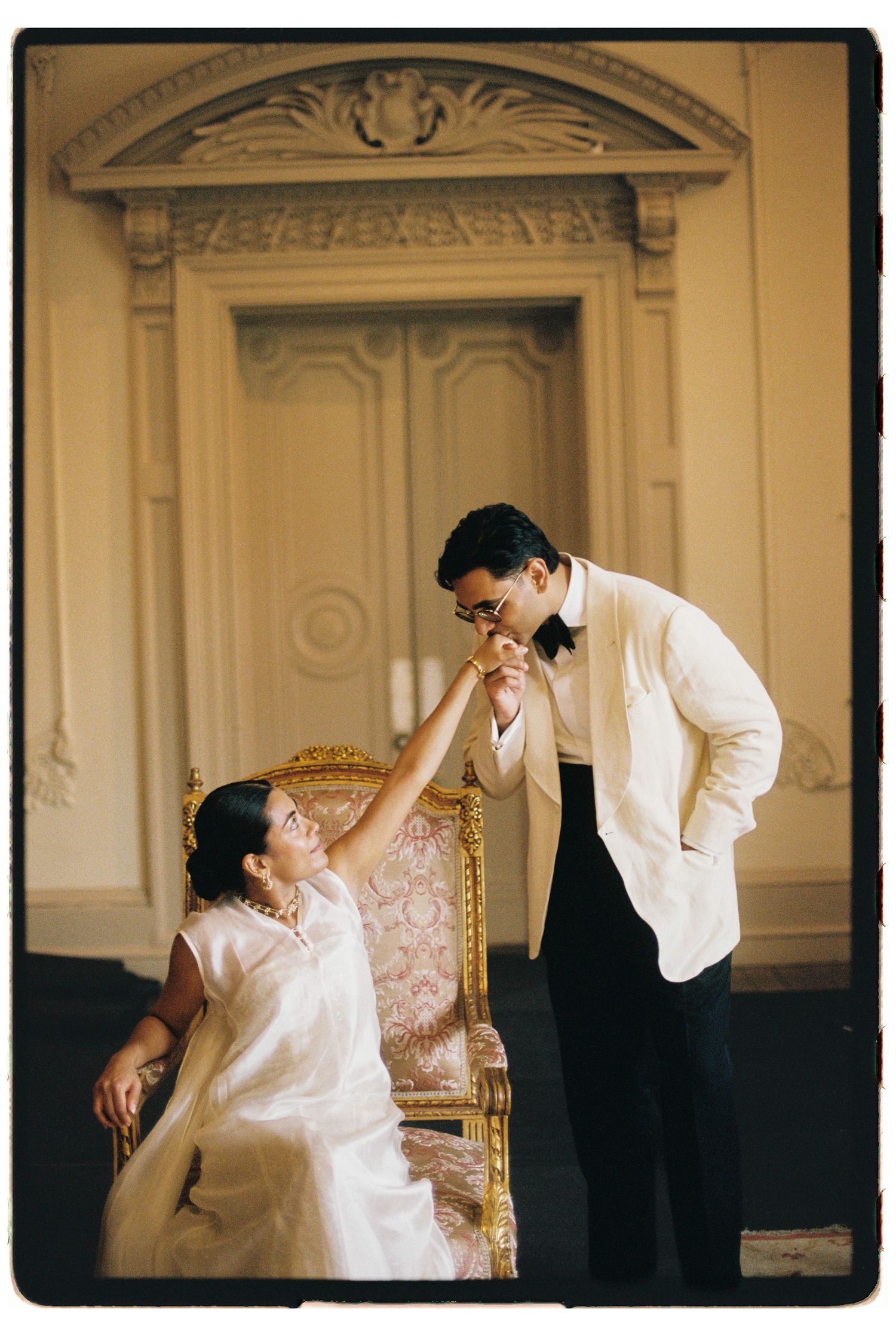 Groom in white tuxedo kisses bride's hand as she sits on ornate gold chair in elegant venue.