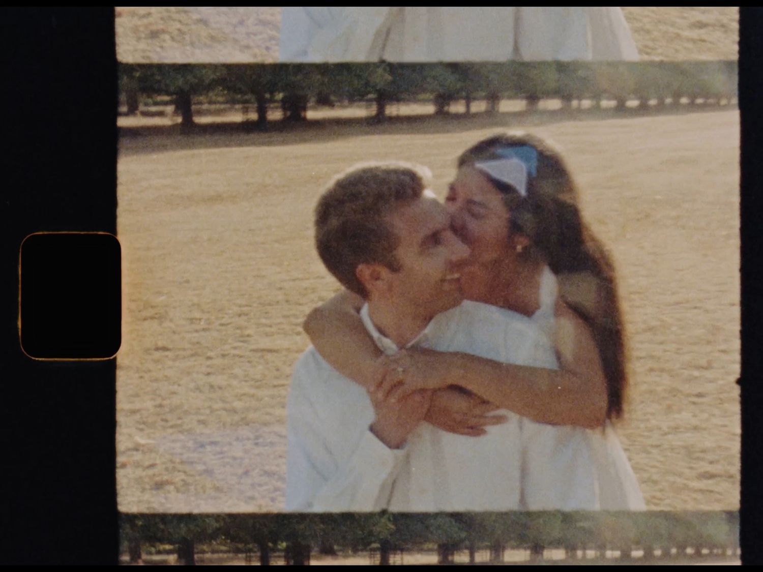 Vintage film photo of a couple embracing outdoors, woman in white dress hugging man from behind in sunny field.
