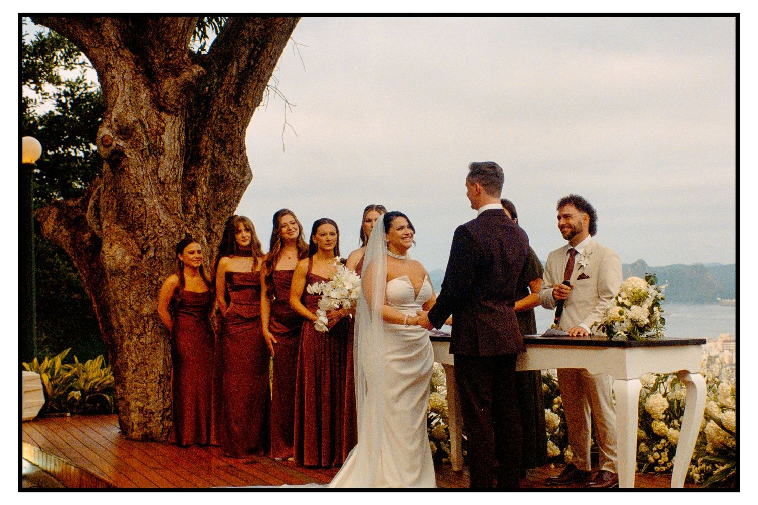 Bride and groom exchanging vows with bridesmaids in burgundy dresses under a large tree by water.