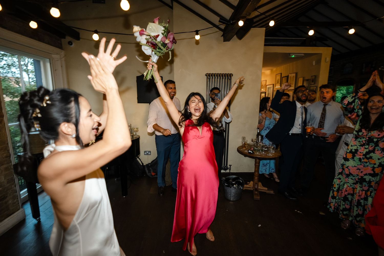 Bride tossing wedding bouquet to excited guest in red dress at lively reception party.