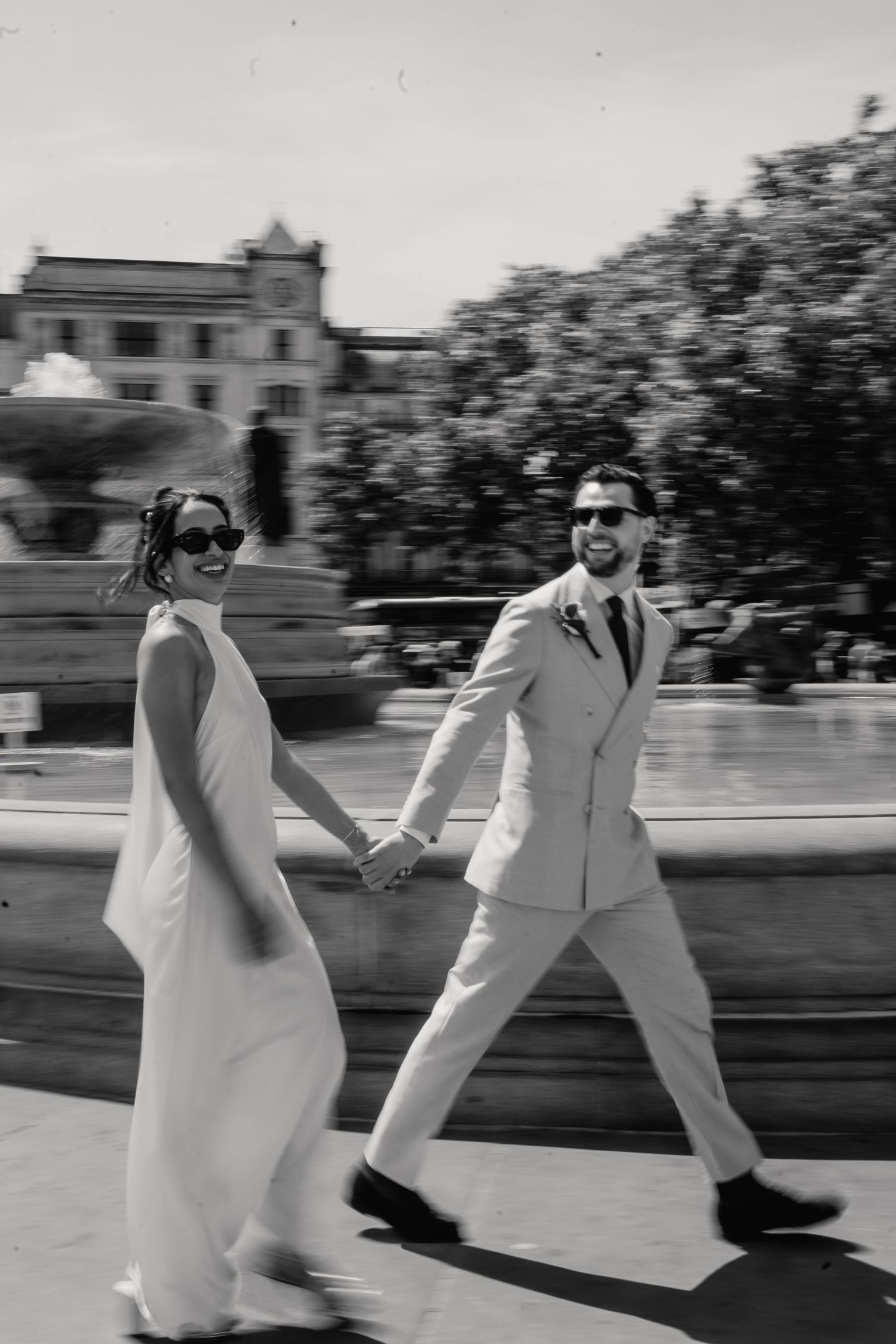 Black and white motion blur photo of newlyweds walking hand in hand past Trafalgar Square fountain.