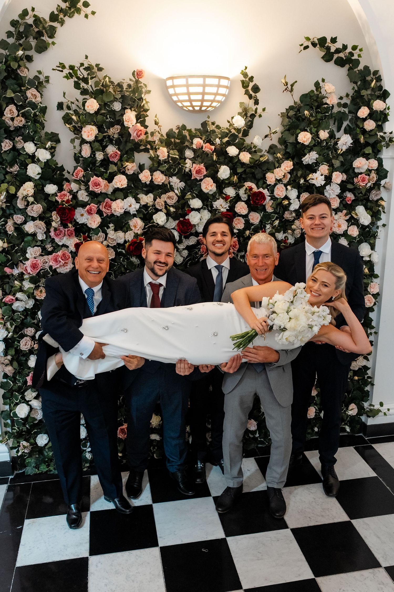 Groomsmen lifting smiling bride in white gown before floral wall with pink and white roses.