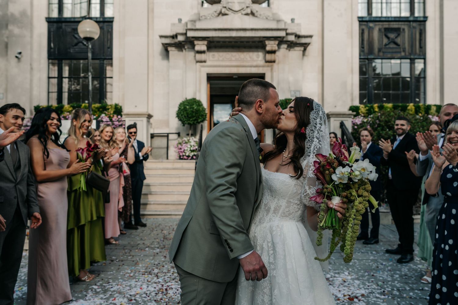 Newlyweds share a kiss outside a grand building surrounded by cheering guests and colorful bouquets.