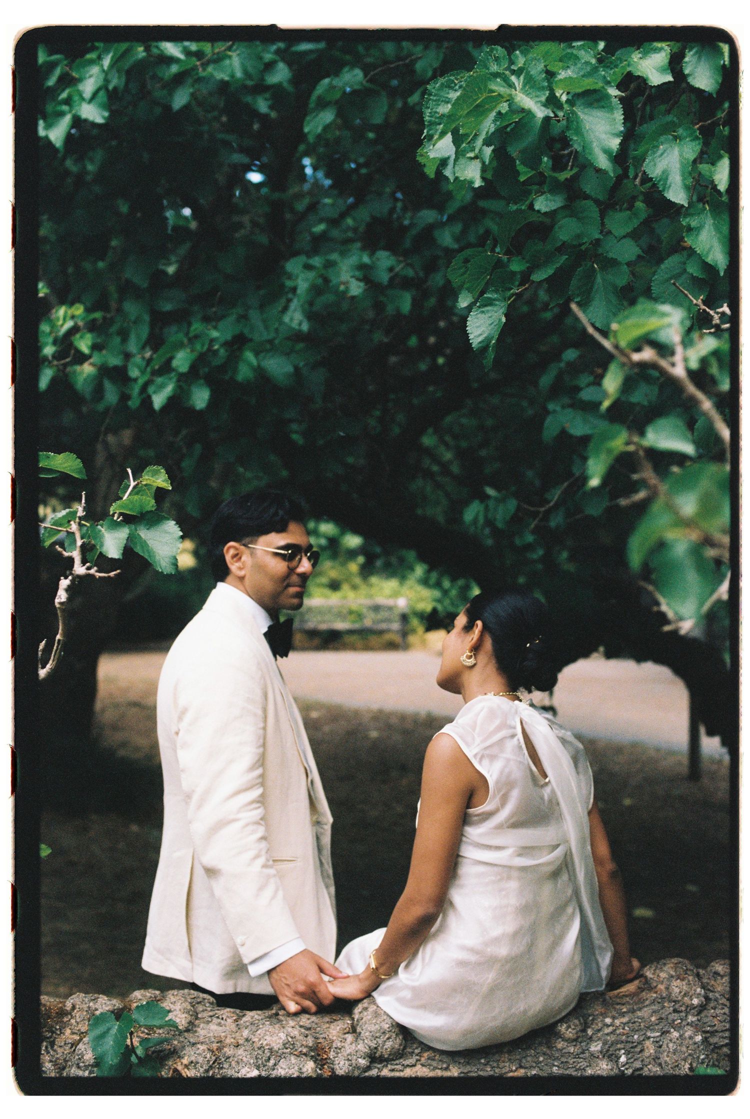 Couple in white attire holding hands under lush green trees in a romantic outdoor setting.