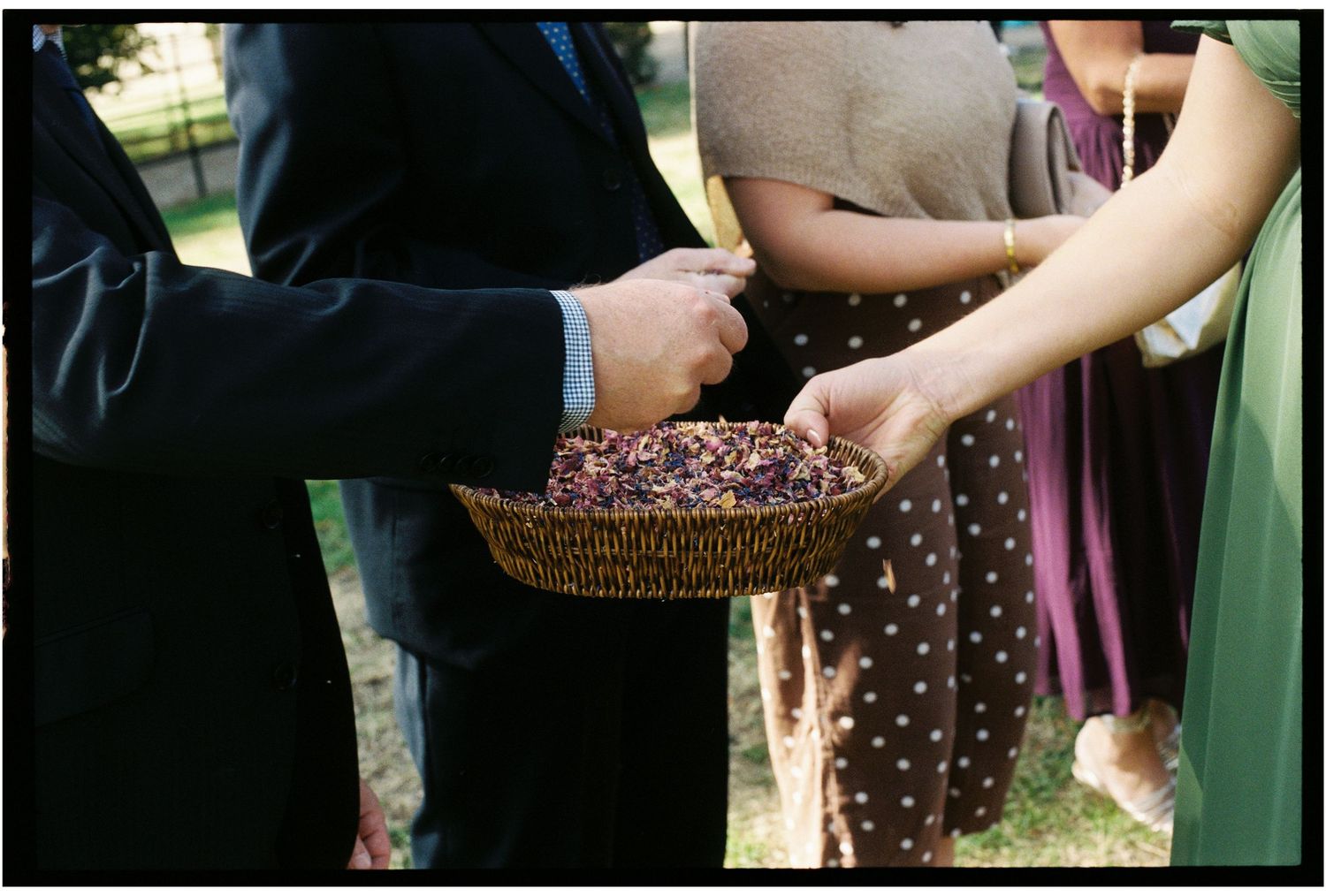 Guests hold a wicker basket filled with dried flower petals at an outdoor wedding ceremony.